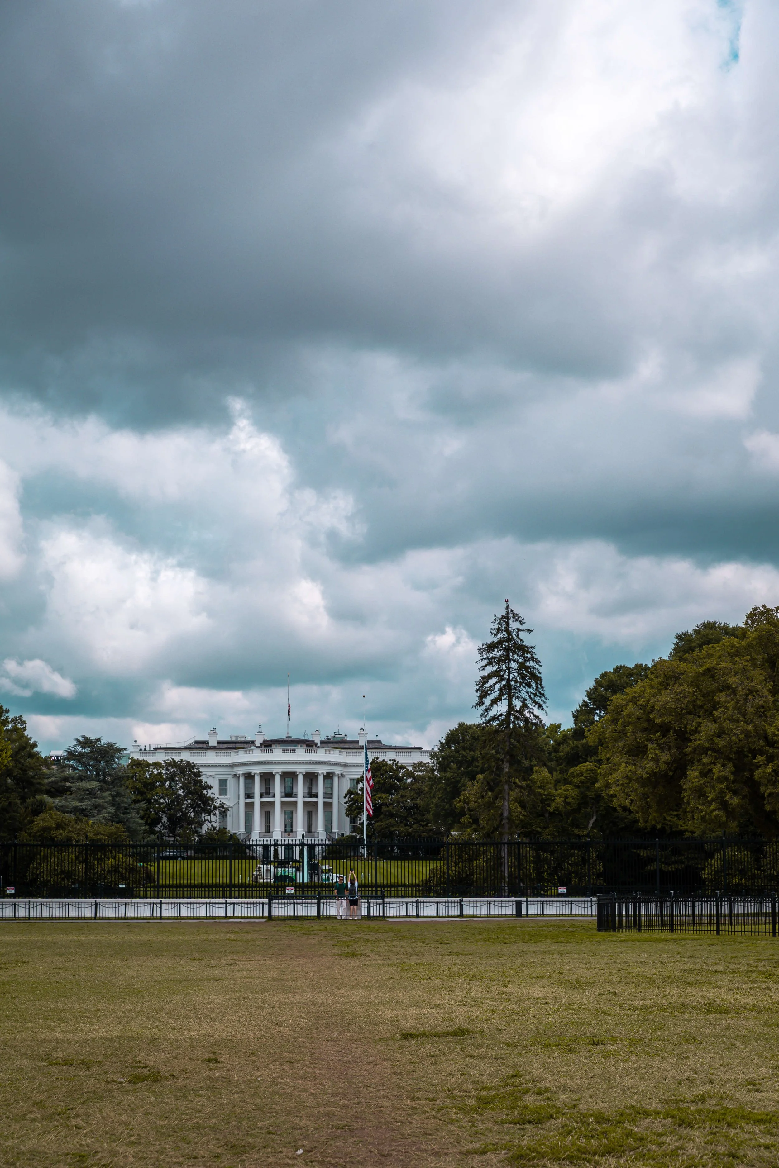 The White House under a cloudy sky, with a fenced lawn and two people standing in front of the fence.