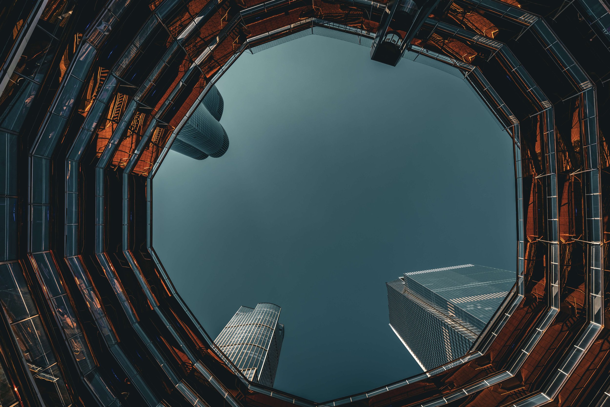 Looking up at a modern glass skyscraper courtyard from the ground, with tall buildings surrounding a clear blue sky