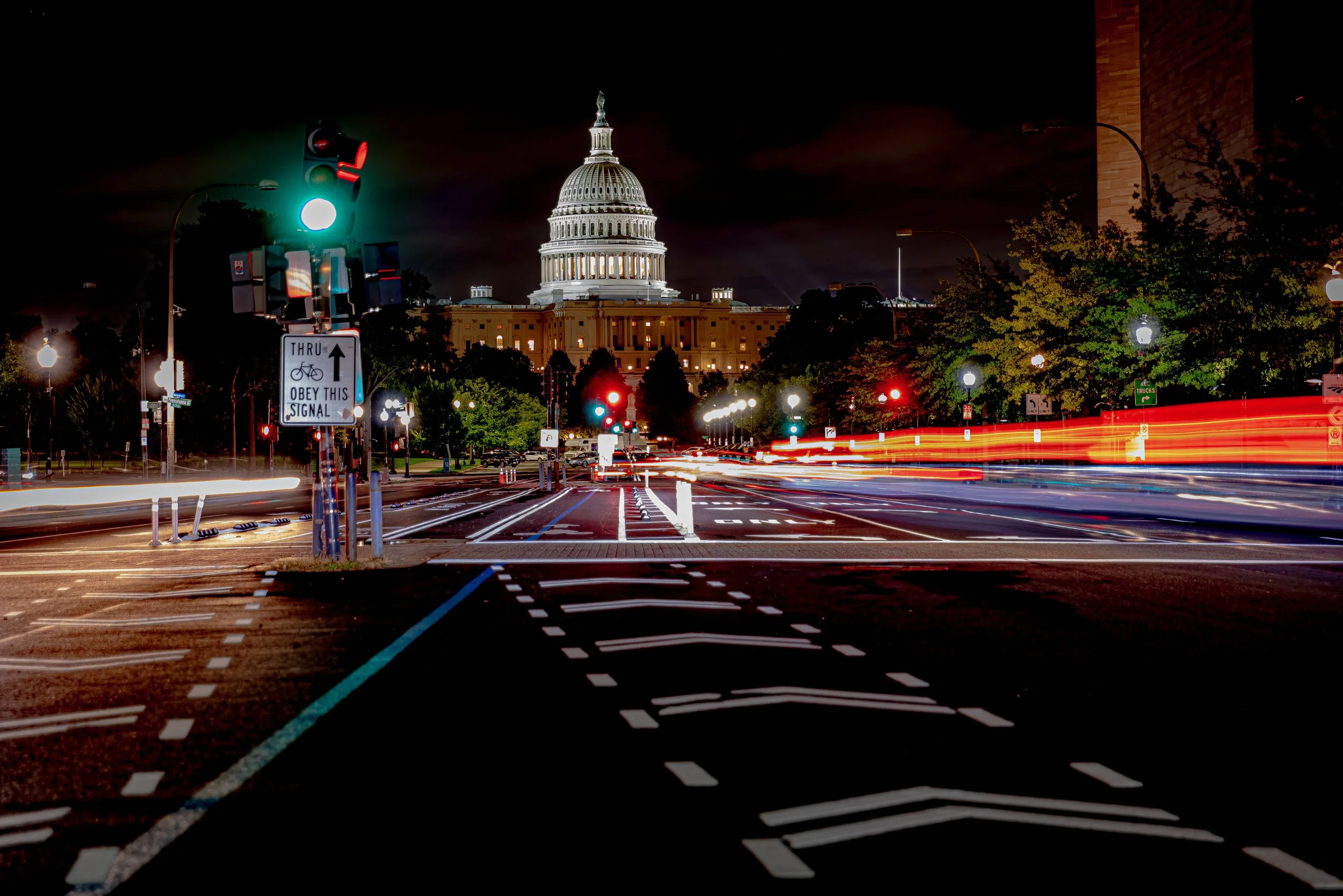 Nighttime city street with light trails from moving vehicles, traffic lights, and the U.S. Capitol building illuminated in the background.