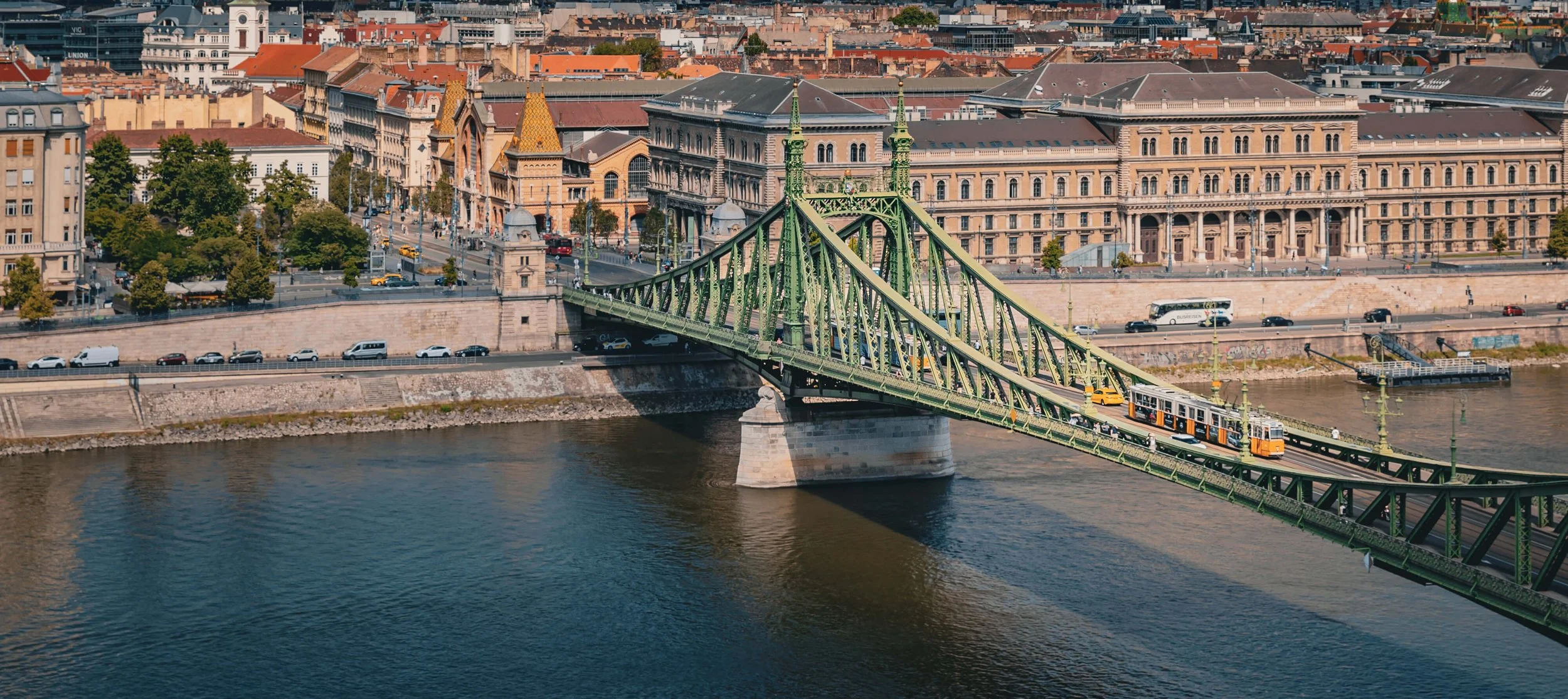 A cityscape with a green suspension bridge over a river, a vintage tram crossing the bridge, and historic buildings along the riverbank.