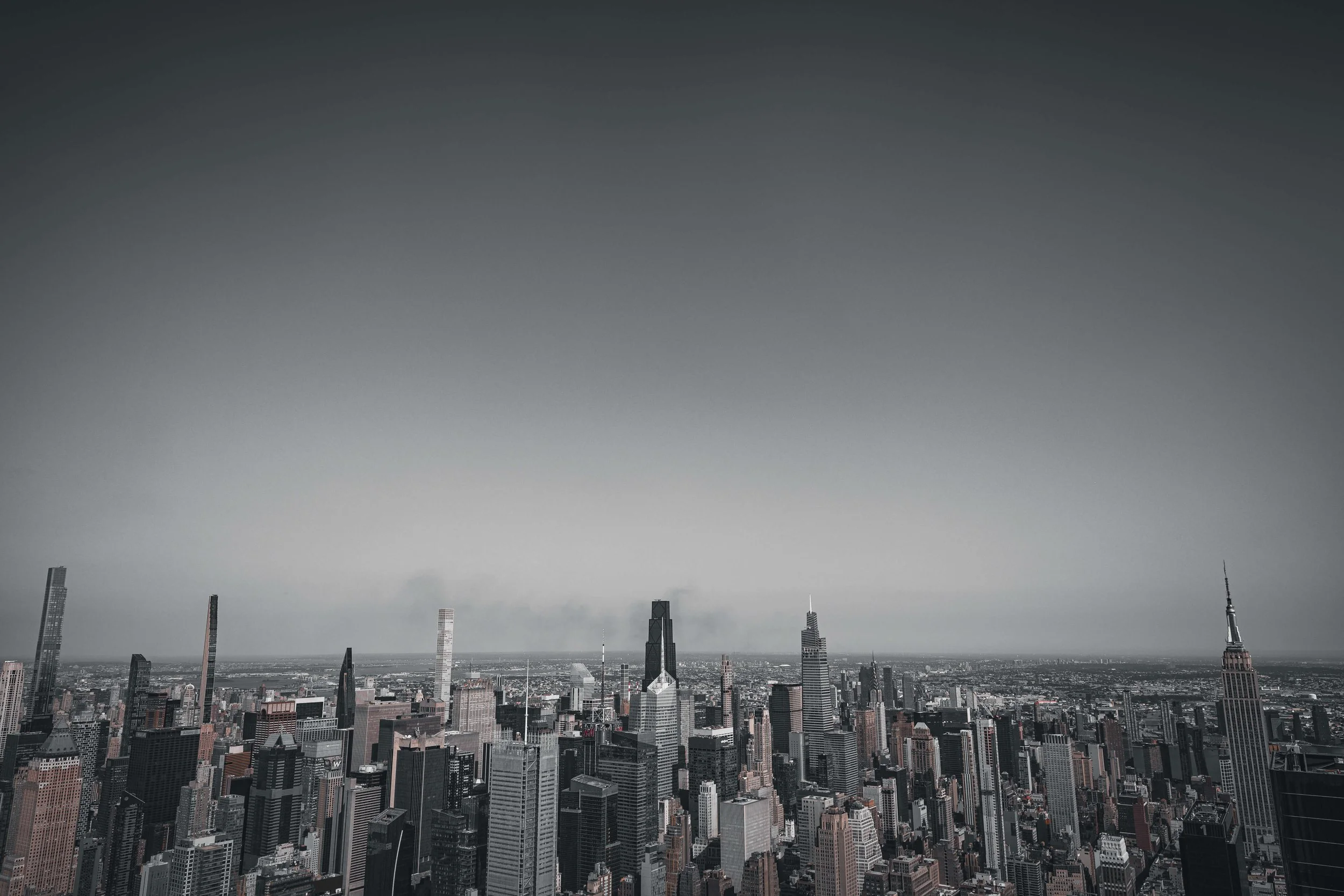A black-and-white aerial view of a city skyline with numerous skyscrapers, including the Empire State Building on the right, under a clear sky.