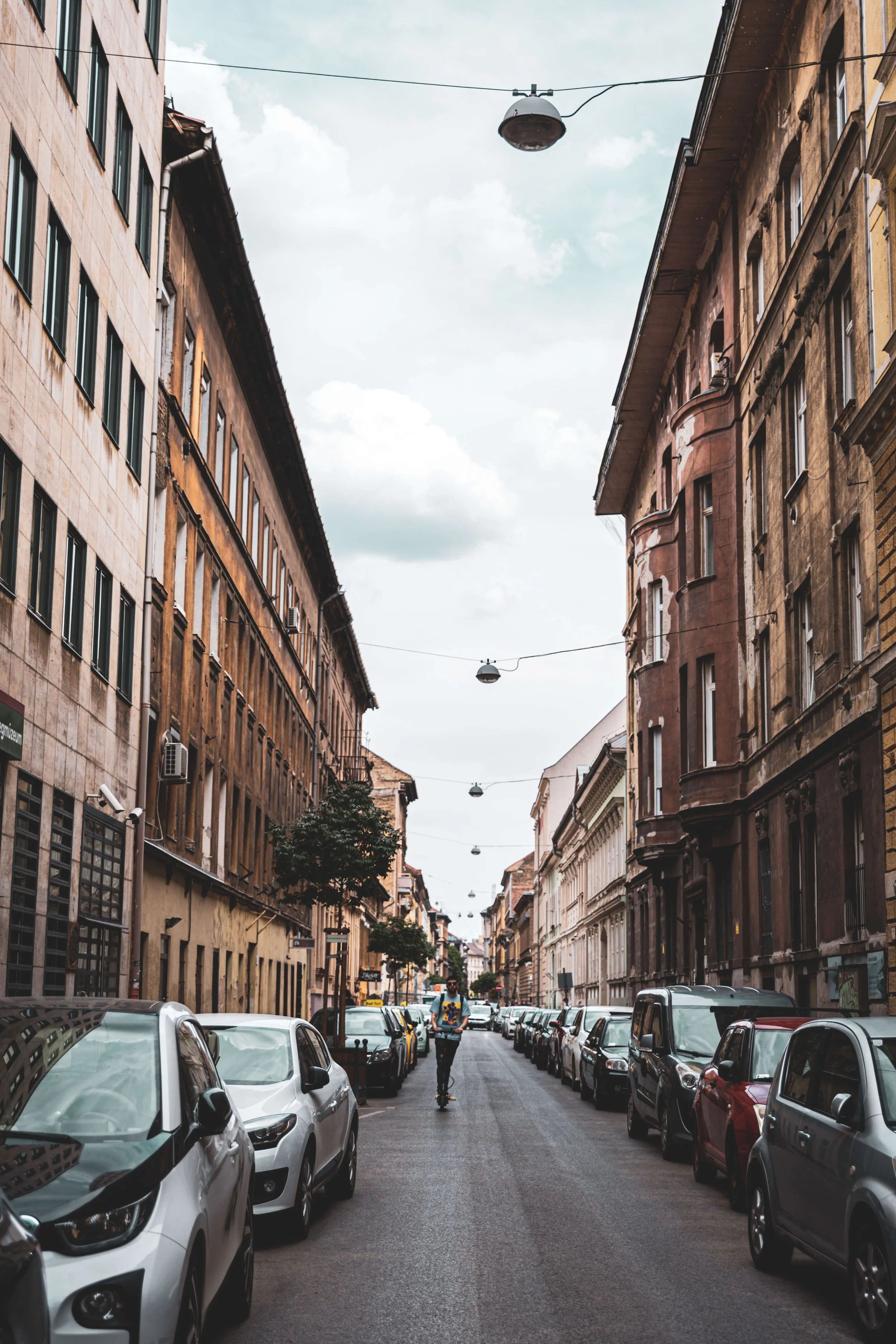 A city street scene with parked cars on both sides, historic buildings with multiple windows lining the street, a man riding a scooter in the middle, trees along the sidewalk, and a cloudy sky overhead.