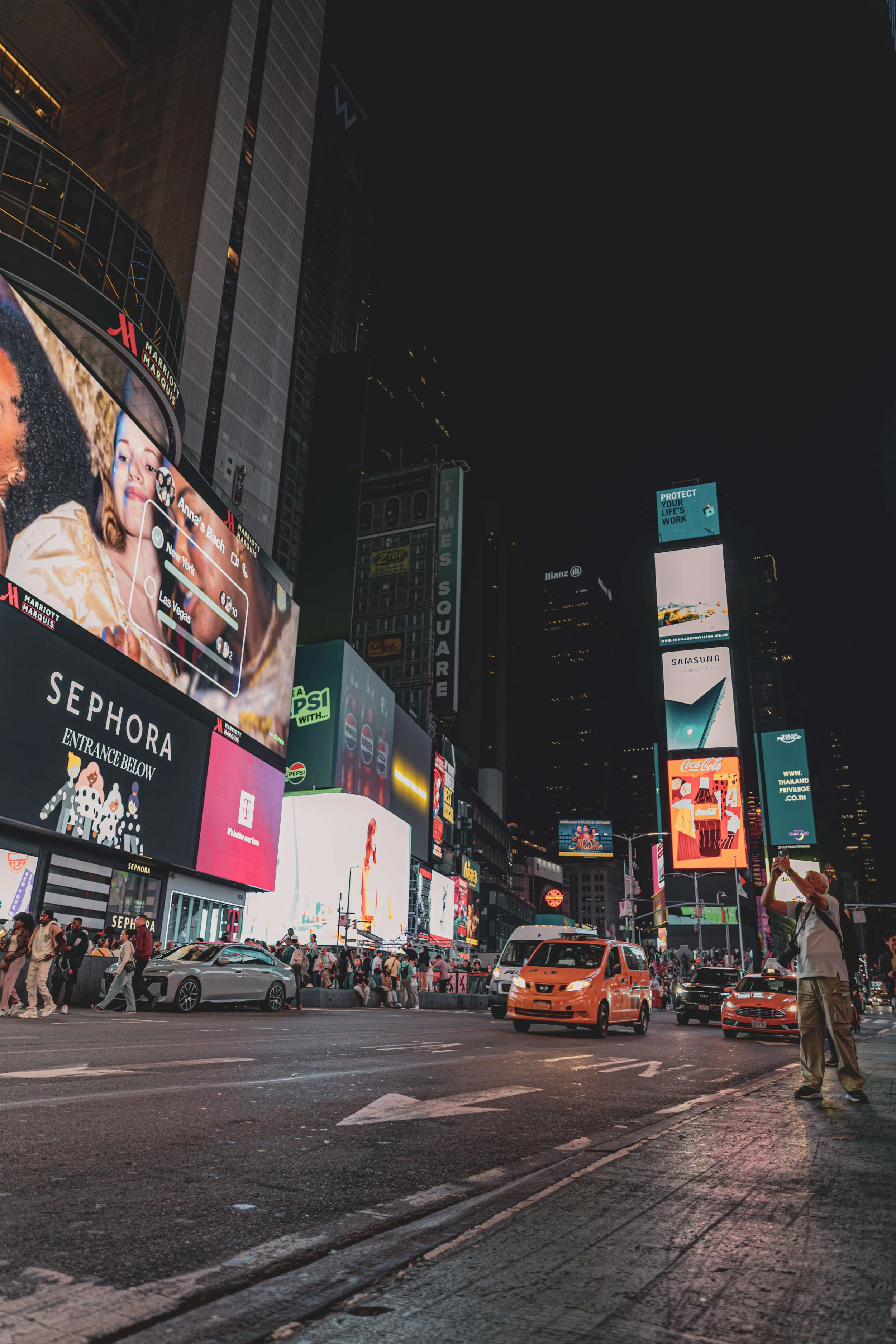 Nighttime scene in Times Square, New York City, with bright digital billboards, illuminated advertisements, people walking on the sidewalk, and vehicles on the road.