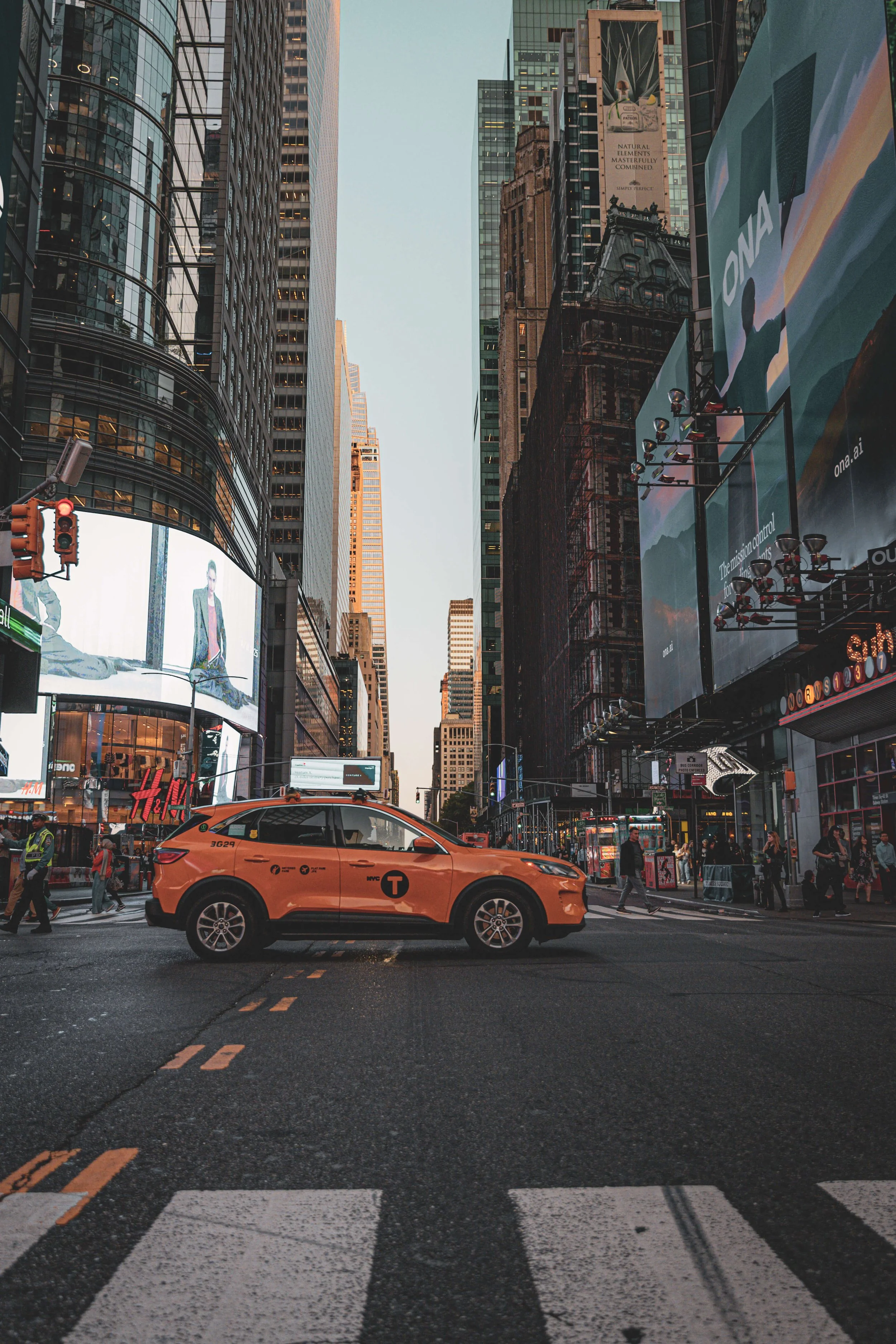 A bright orange NYC taxi cab crossing a busy intersection in Times Square, surrounded by illuminated billboards and tall skyscrapers.