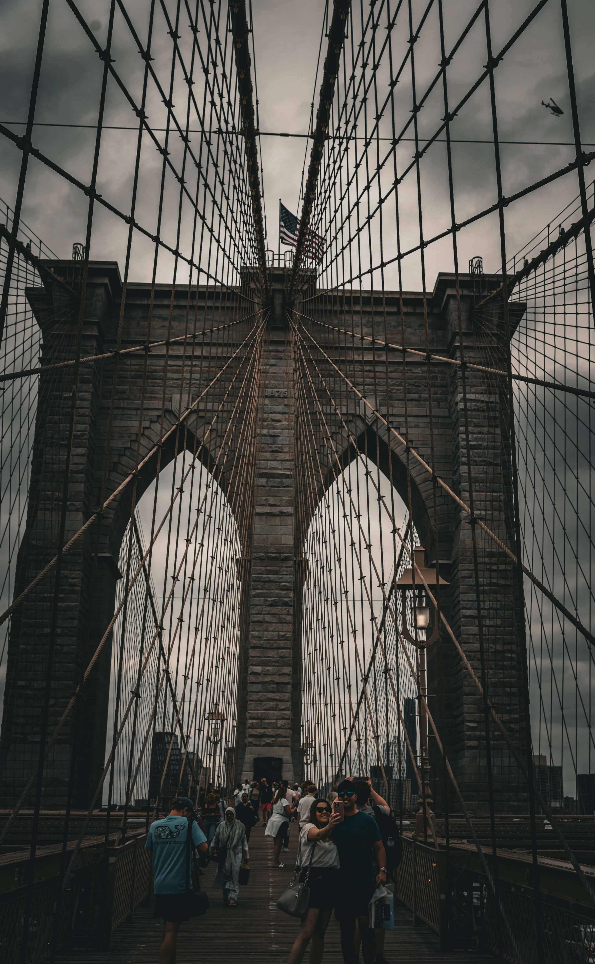 People walking on the Brooklyn Bridge with the Manhattan skyline in the background on a cloudy day, view from below the bridge's cables.