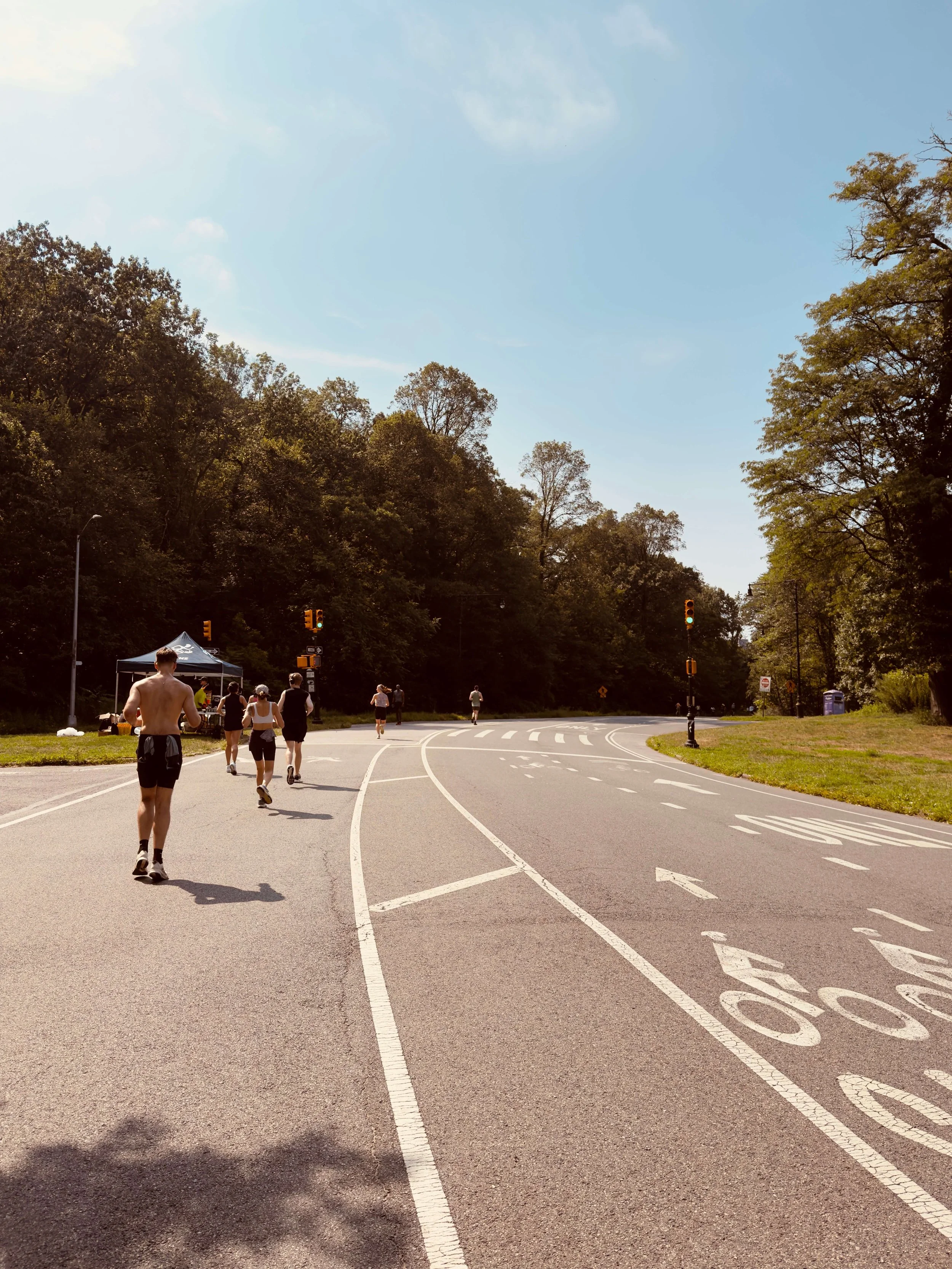 People running on a paved pathway through a park surrounded by trees, with traffic signals and a tent in the background on a sunny day.