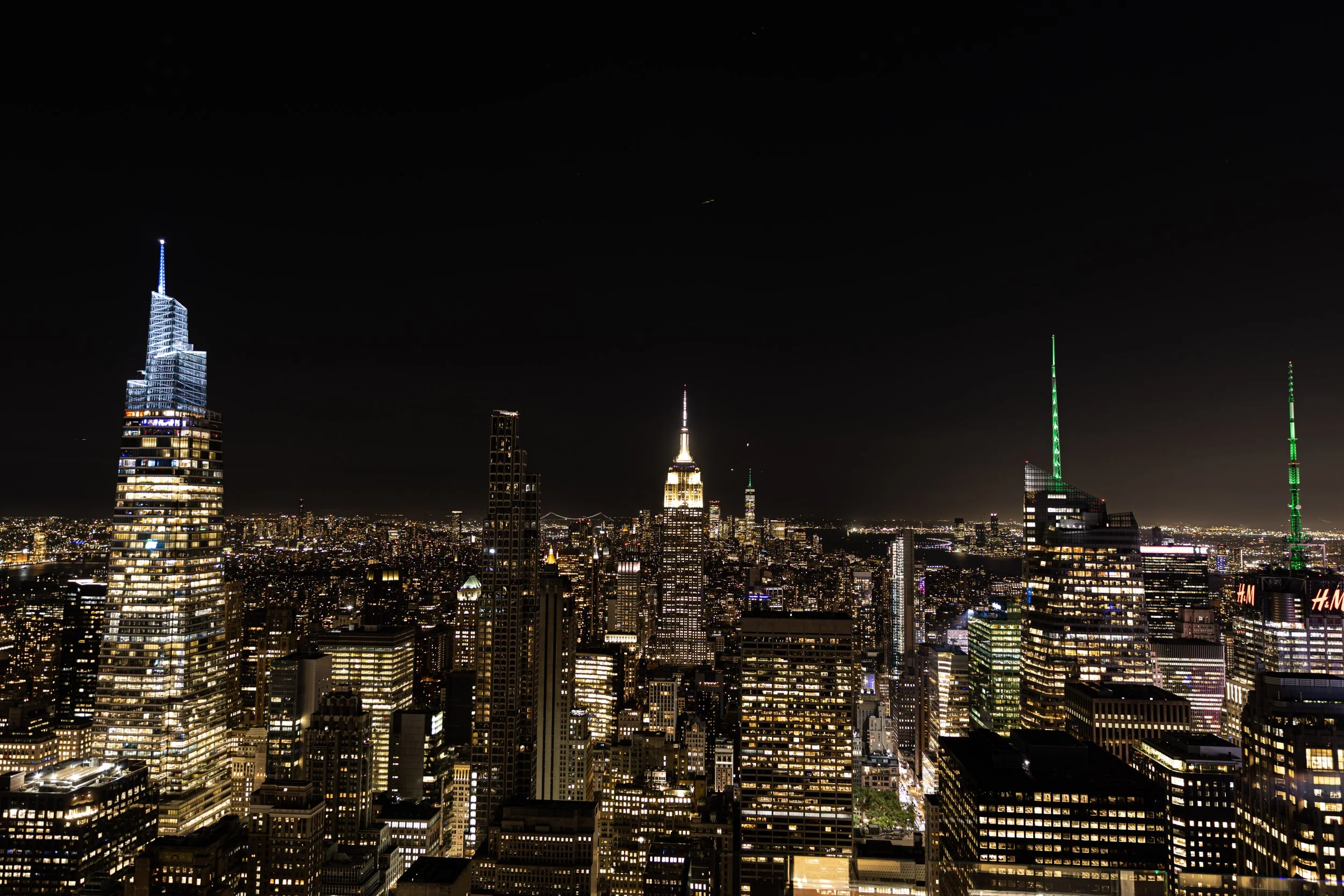 Night skyline of New York City with illuminated skyscrapers including the Empire State Building, surrounded by city lights and a dark sky.
