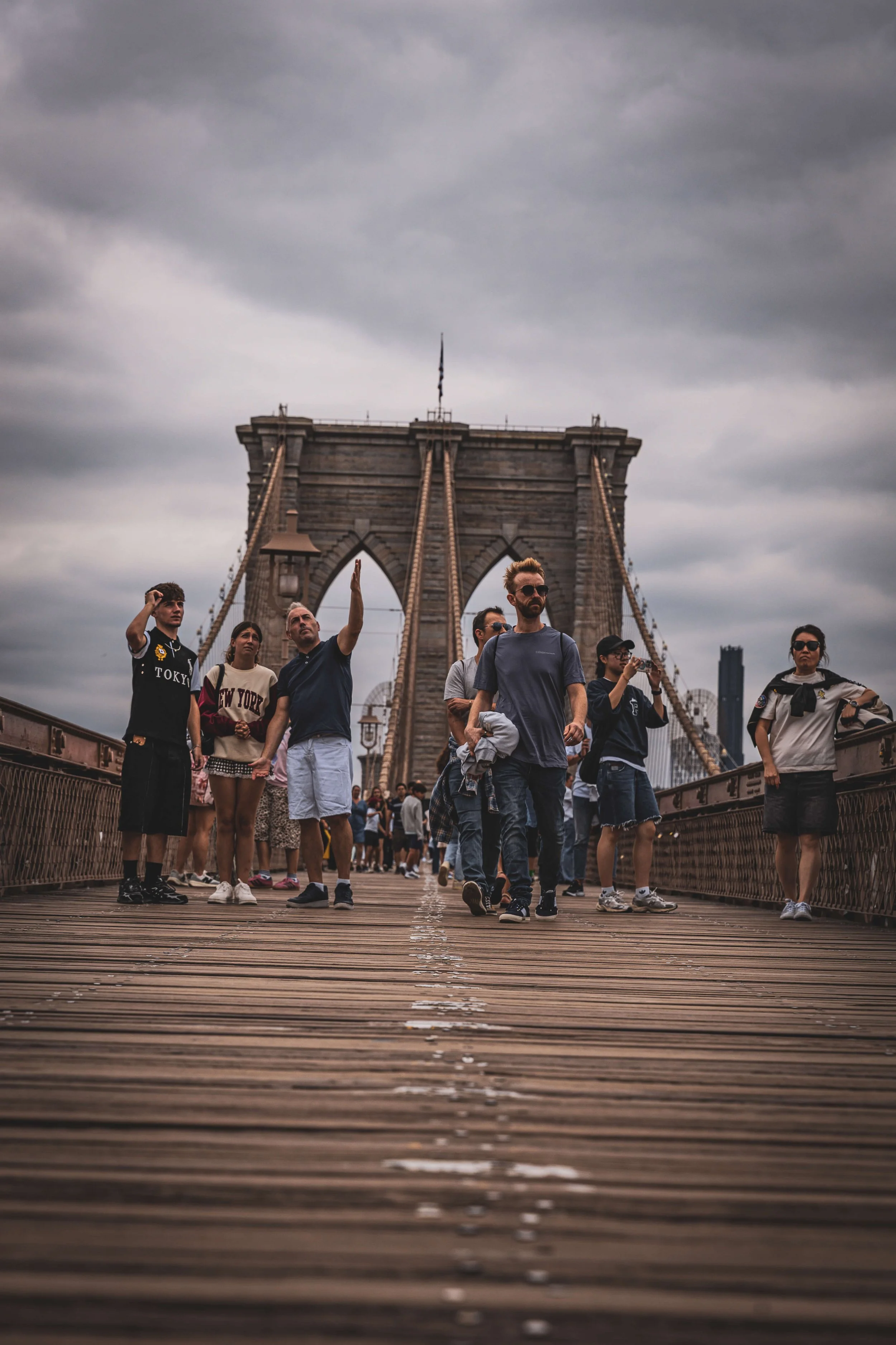 Tourists walking on the Brooklyn Bridge in New York City under a cloudy sky.