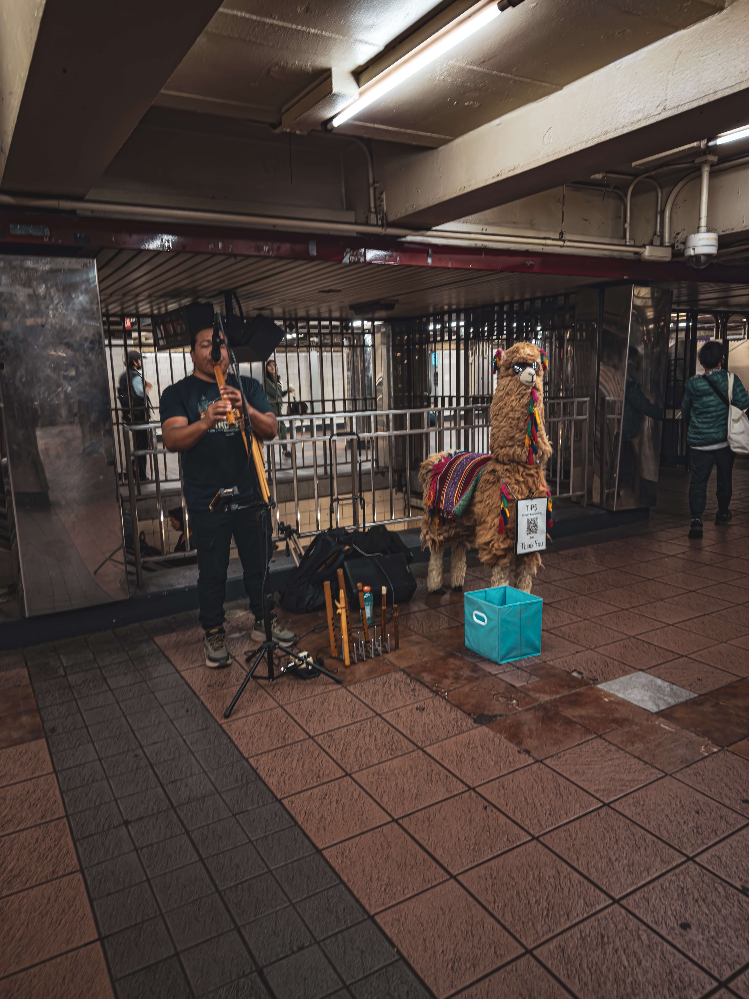 A street performer playing a flute in front of a llama costume with colorful decorations, set up near an underground station entrance.