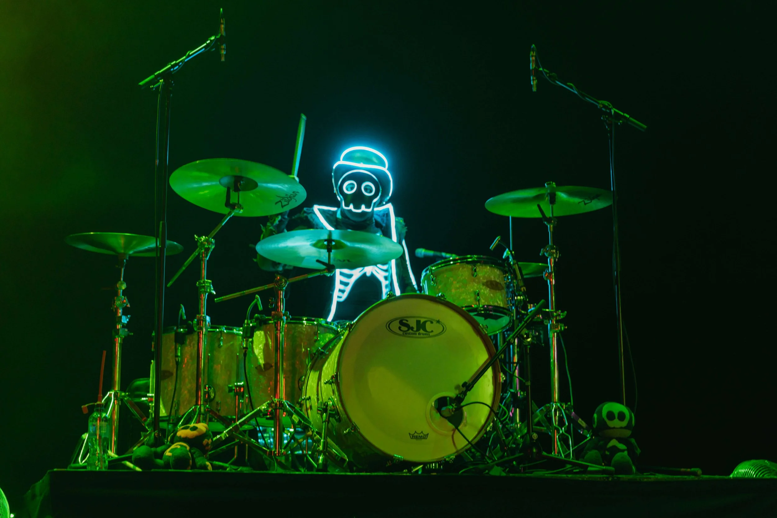 A drummer's drum set illuminated by vibrant green and yellow lights, with plush toys at the base and a neon skeleton figure with a hat in the background.