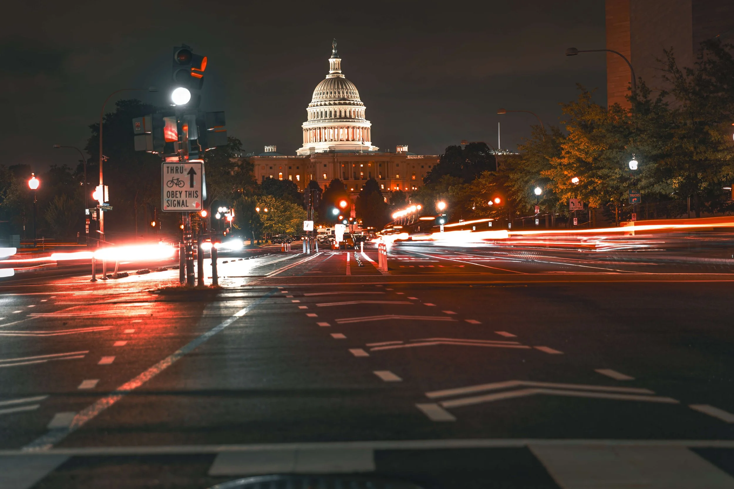 Nighttime view of Washington D.C., showing the U.S. Capitol building illuminated in the background, with light trails from moving vehicles on the street in the foreground.