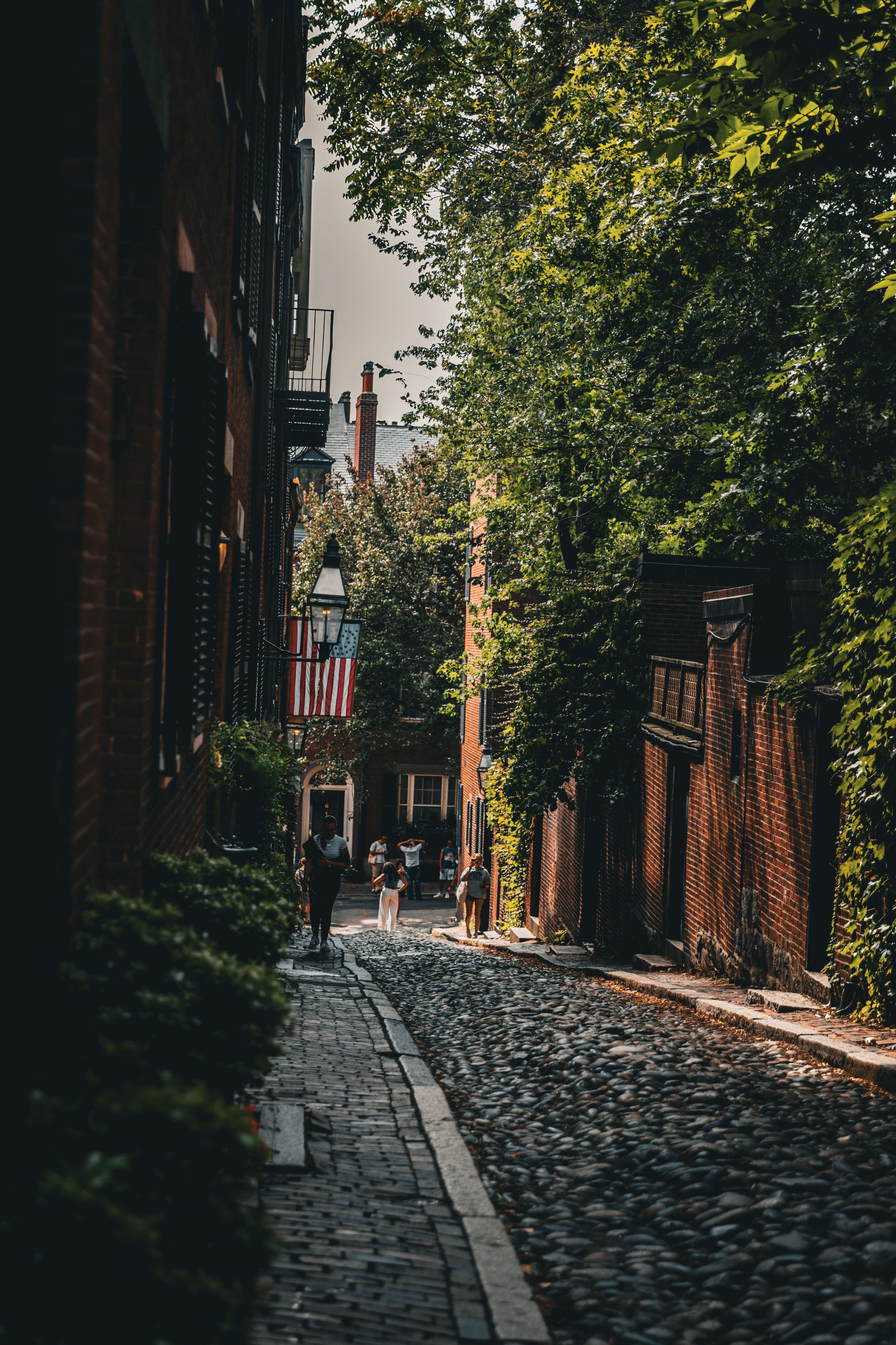 A cobblestone street lined with brick buildings and lush green trees, with several people walking and standing along the sidewalk.
