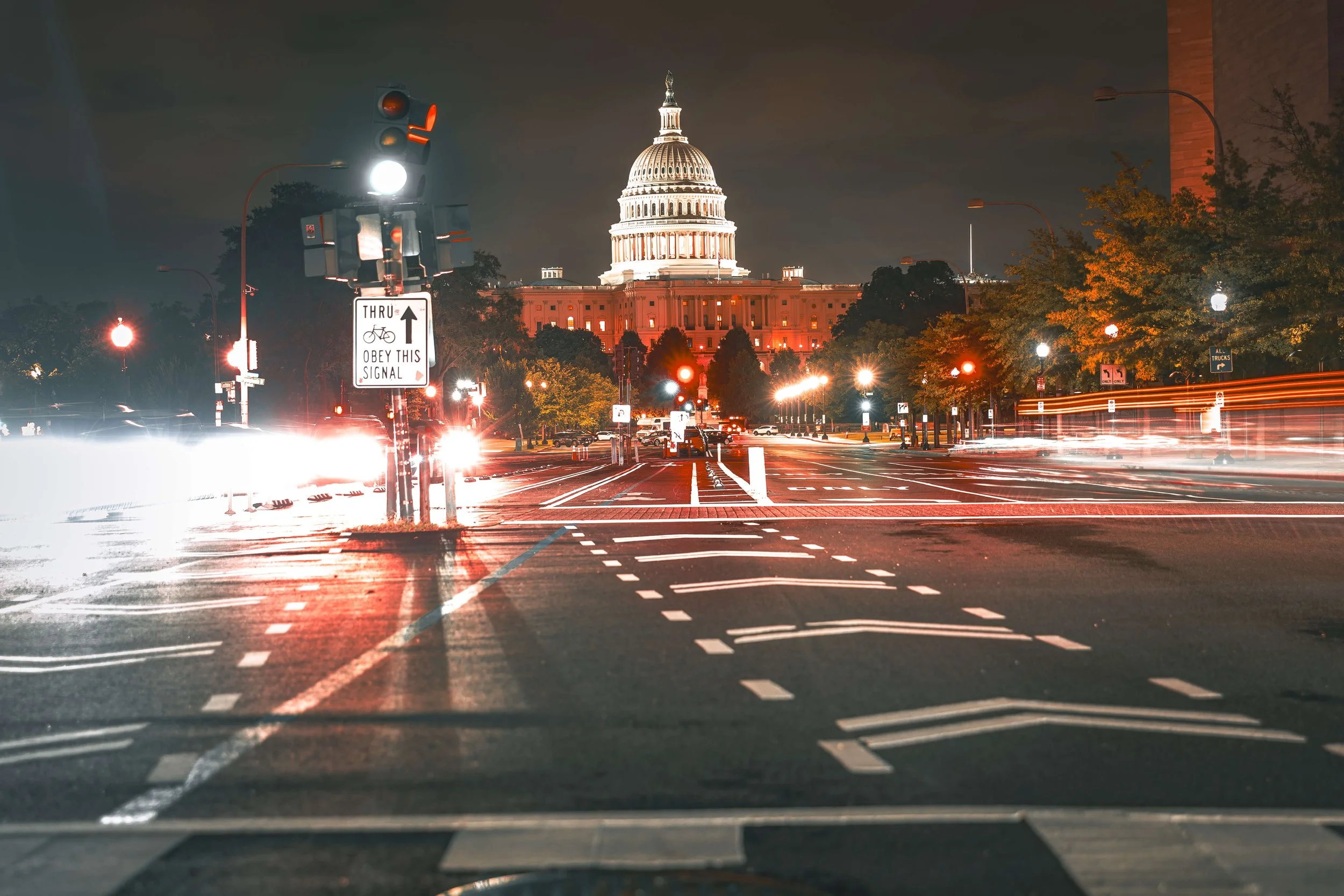 Nighttime view of the US Capitol building in Washington, D.C., with city streets in the foreground showing light trails from moving vehicles and traffic signals.