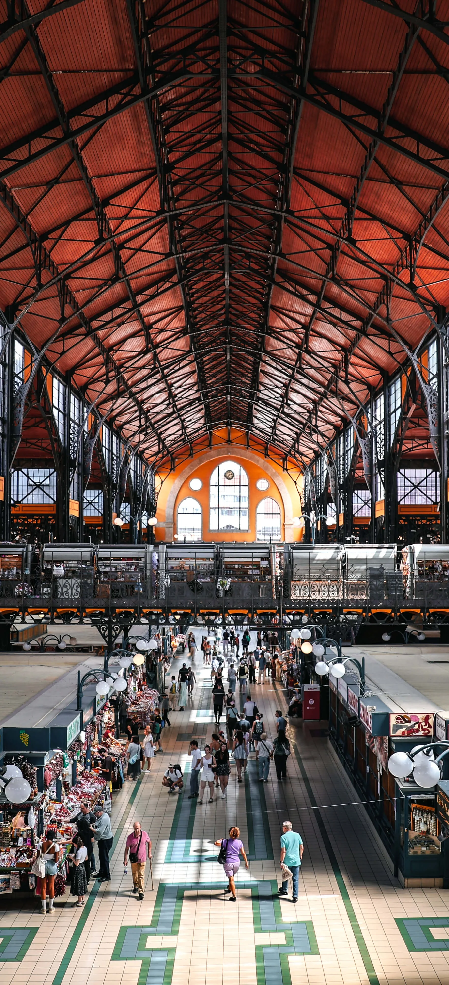 Interior of a historic market hall with a high arched ceiling, large windows, and a bustling marketplace with shoppers and vendors.