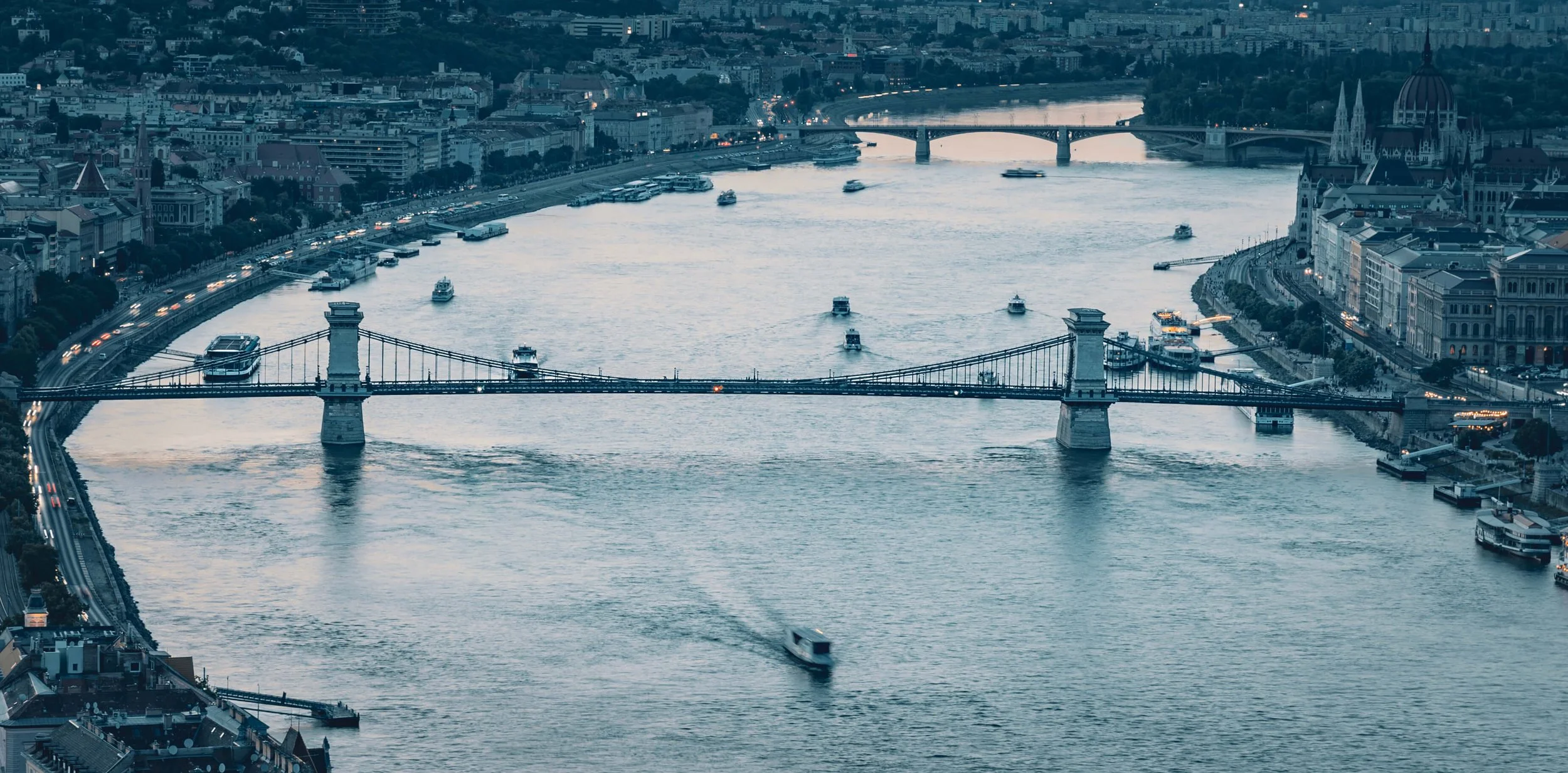 Aerial view of a city river with multiple boats, bridges, and surrounding buildings, including a notable Gothic-style cathedral.