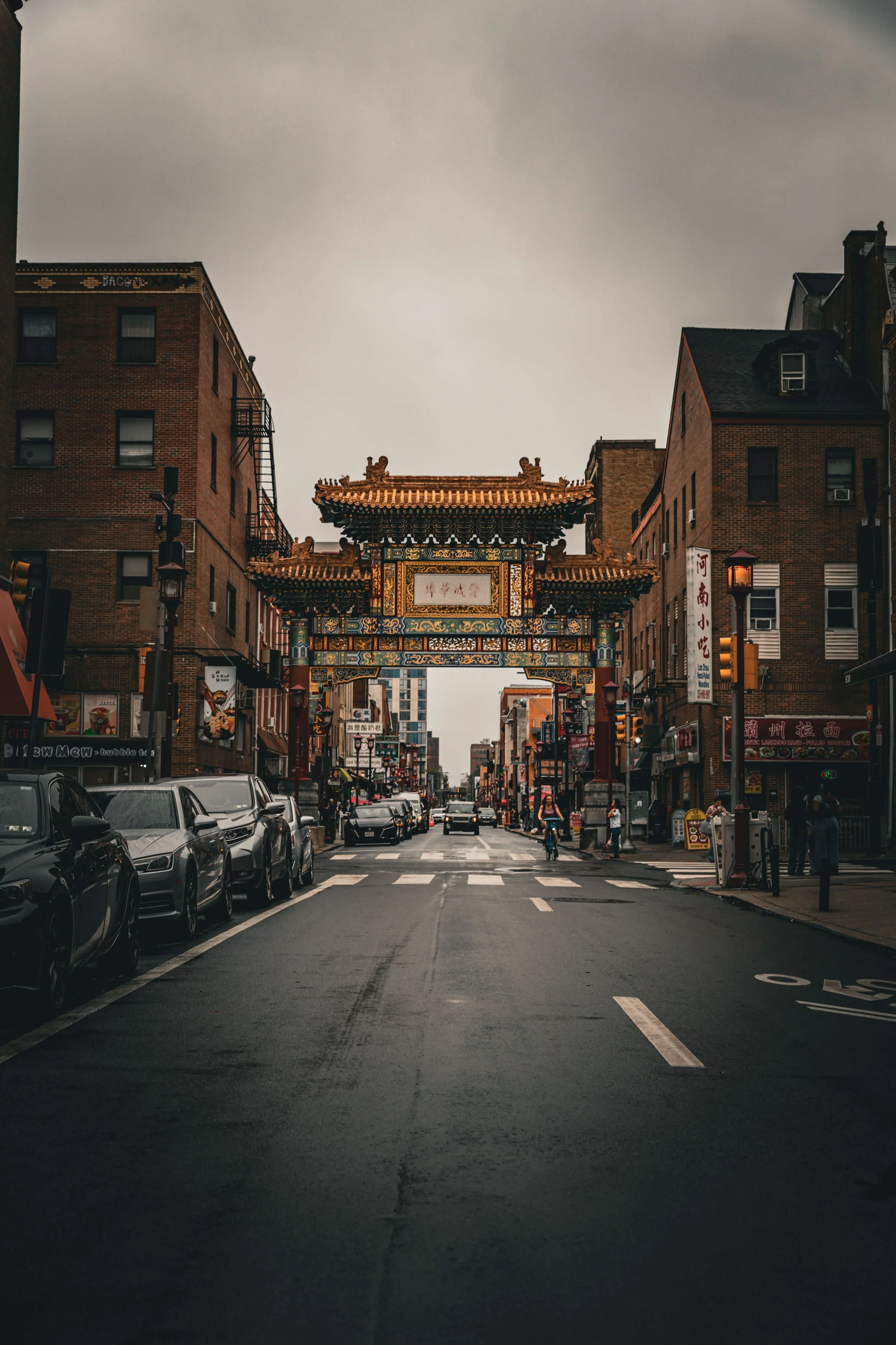 Street scene in Chinatown with traditional Chinese archway, parked cars, pedestrians, and brick buildings on a cloudy day.