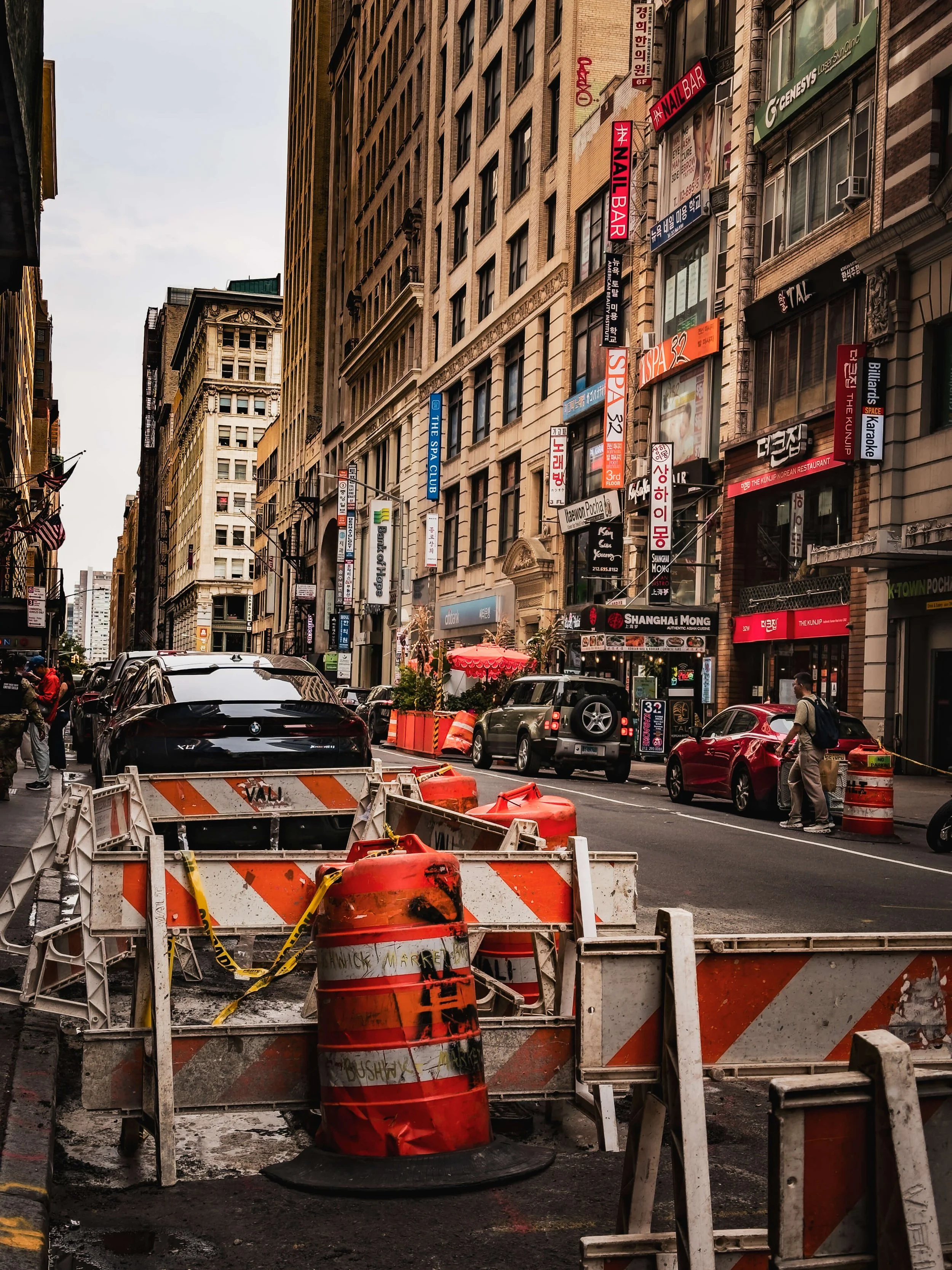 City street with construction barriers and orange cones in the foreground, cars parked along the road, and tall buildings with various signs in the background.