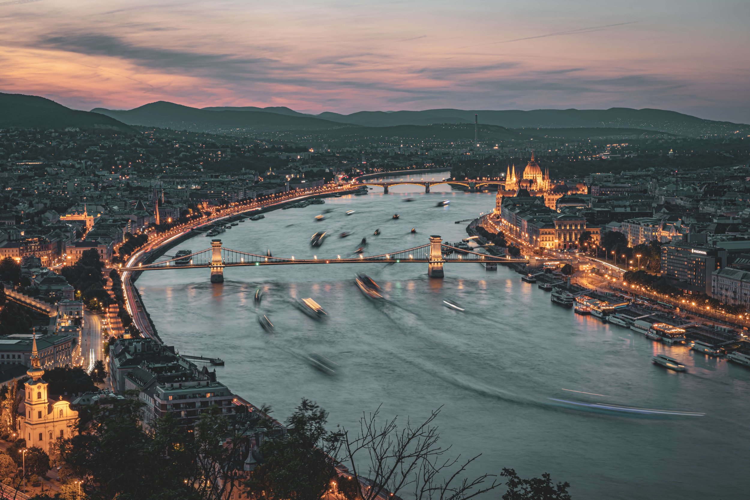 Aerial view of Budapest at dusk showing the illuminated Chain Bridge, the Parliament building, and the Danube River with boats, mountains in the background, and a colorful sky.