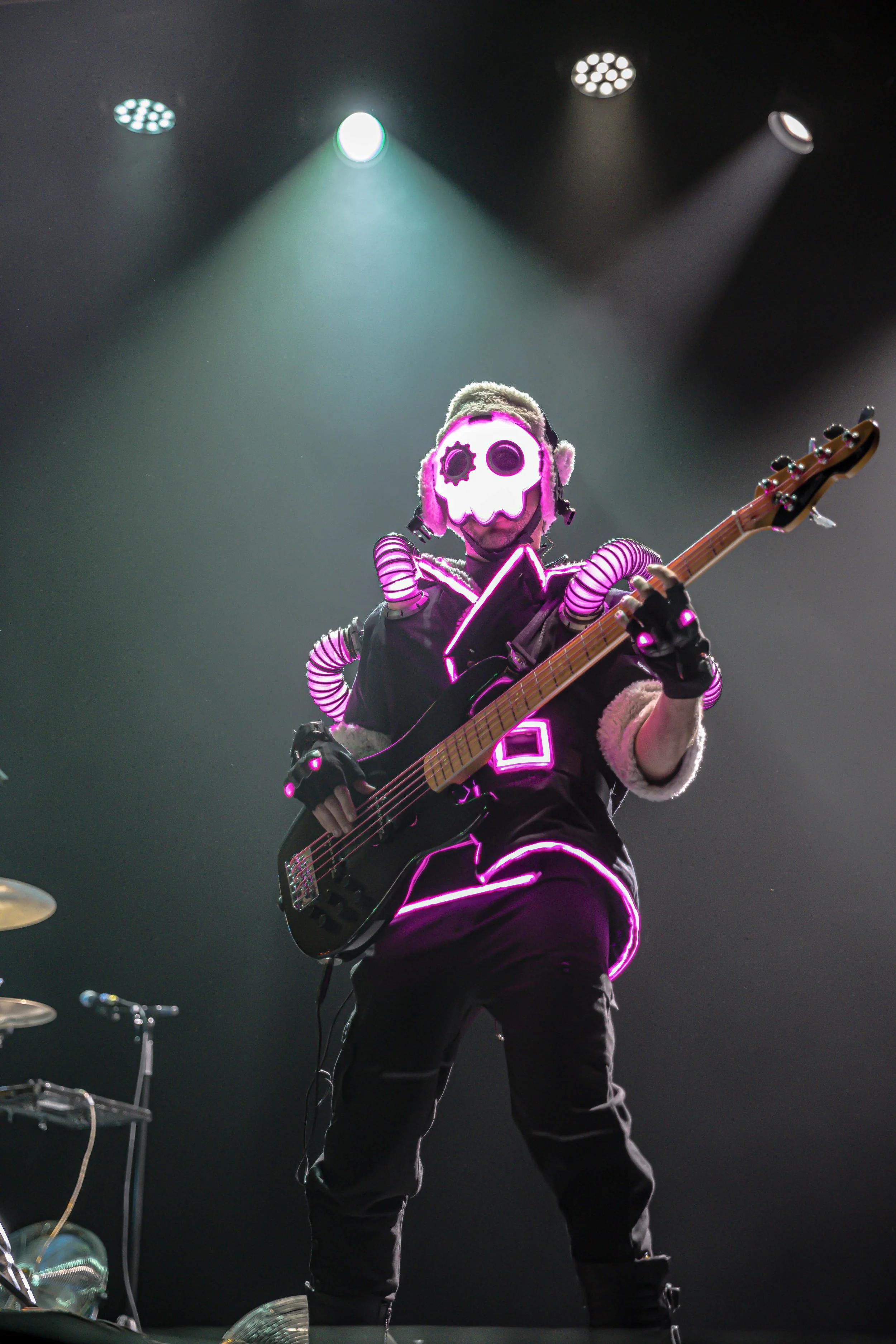 Performer on stage wearing a skull mask with pink neon lights, playing an electric guitar, with dark background and stage lights overhead.