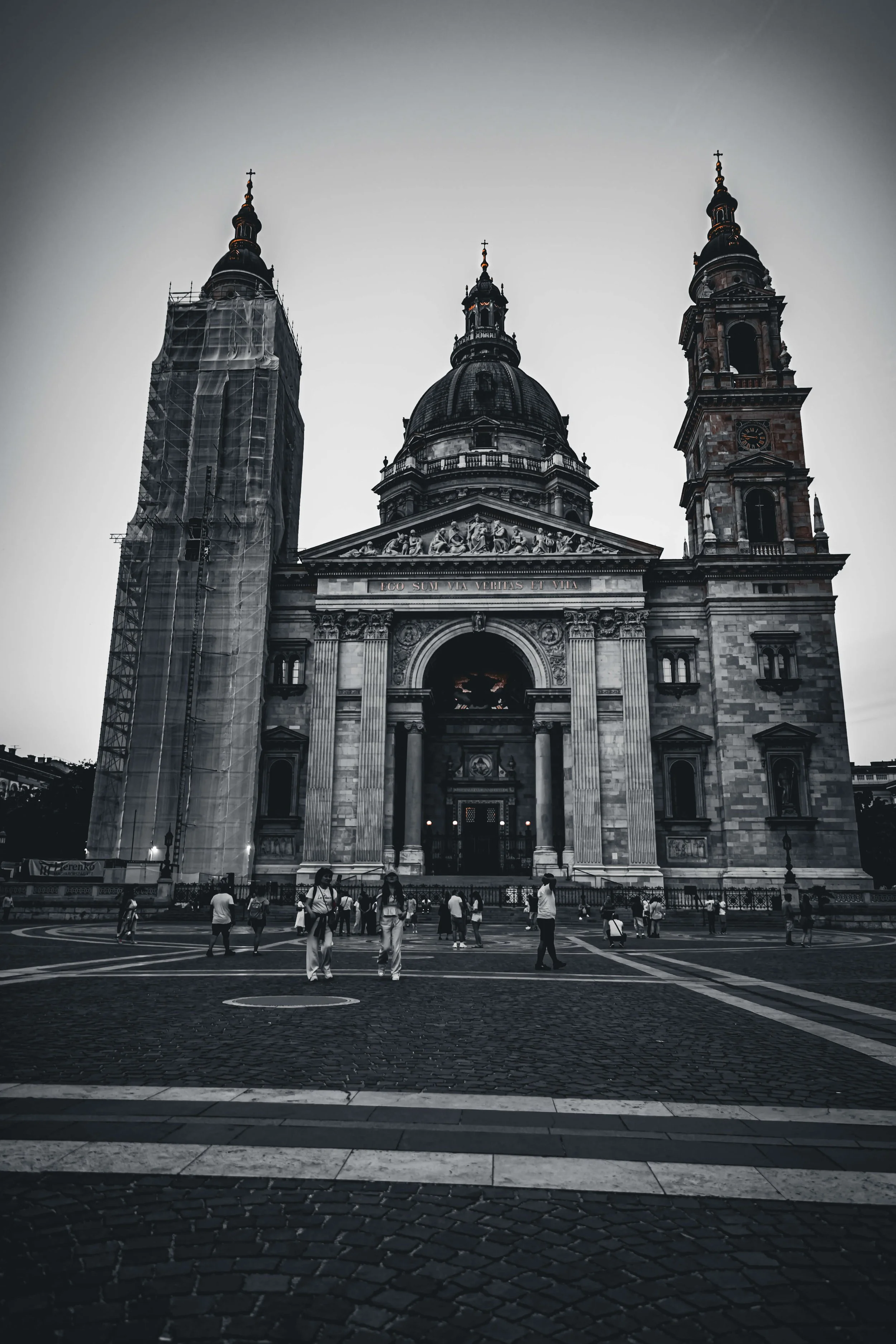 A black and white photo of a large historic church with multiple towers and domes. Several people are gathered and walking in the plaza in front of the church, with some taking photos.