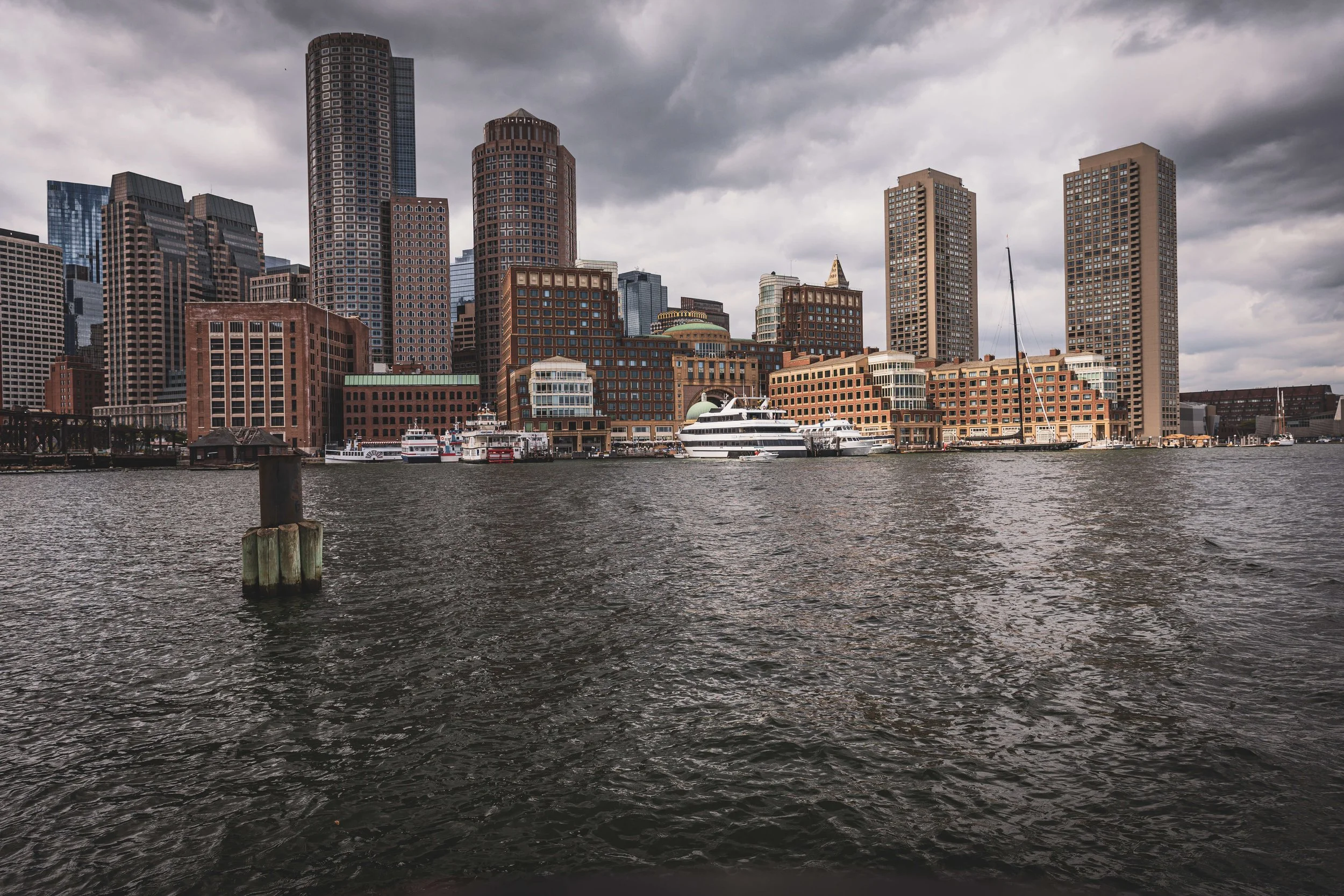 City skyline with tall buildings, boats in the water, and dark cloudy sky.