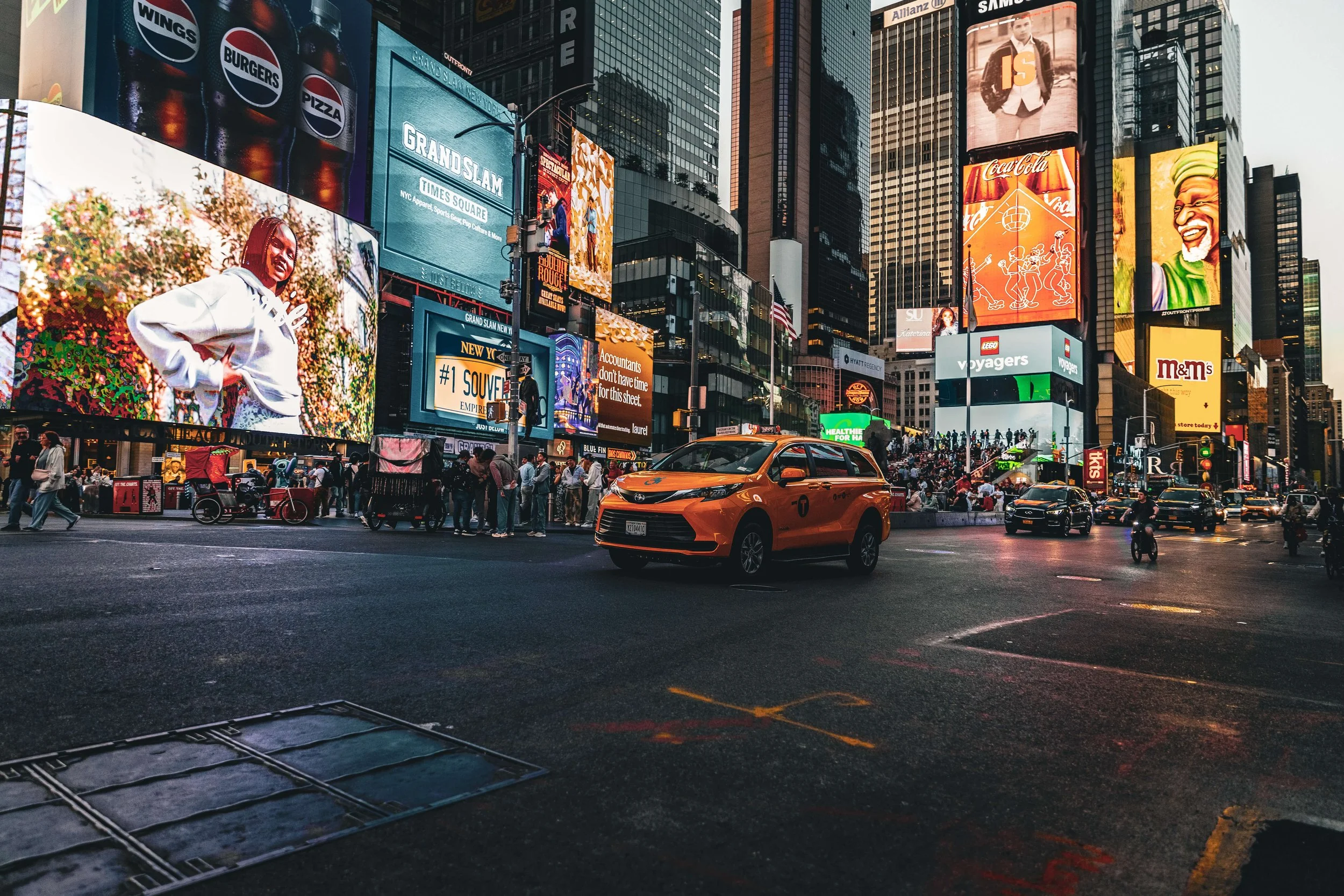Busy city street in Times Square, New York City, with bright digital billboards, crowds of pedestrians, a yellow taxi, and skyscrapers.