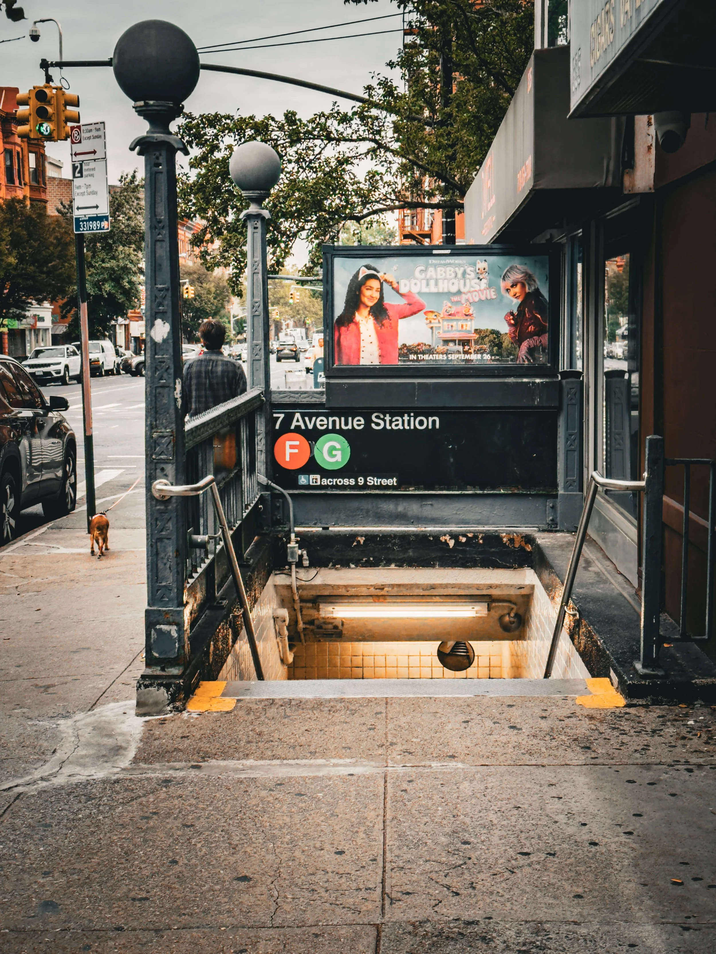 Street entrance to a subway station with steps leading down, located at 7th Avenue Station. Sign showing for subway lines F and G. A man with a dog walks on the sidewalk, and a large advertisement for a movie called Children’s Dollhouse features two 