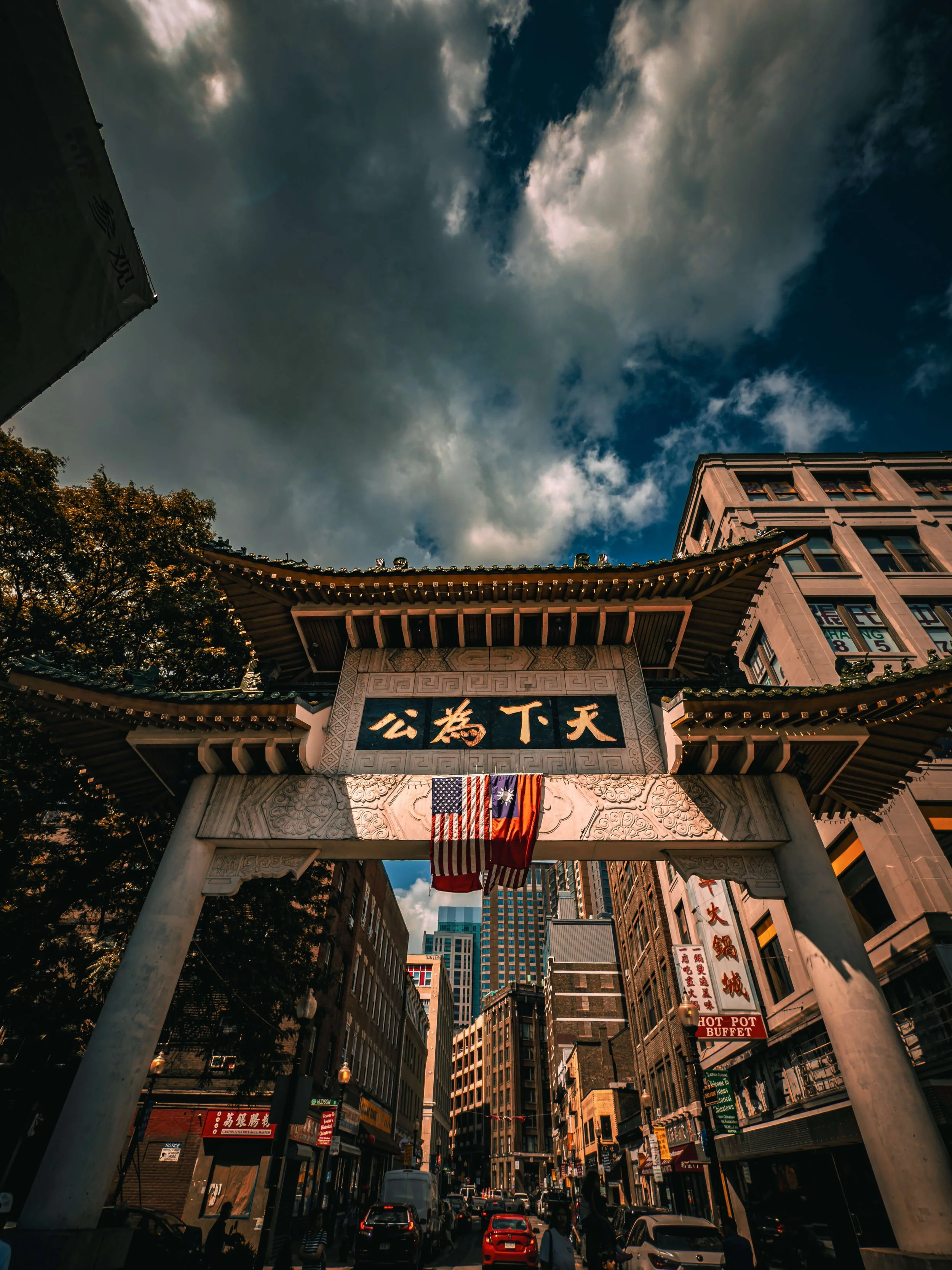 Traditional Chinese archway in a busy city street with buildings, cars, and pedestrians under a partly cloudy sky.