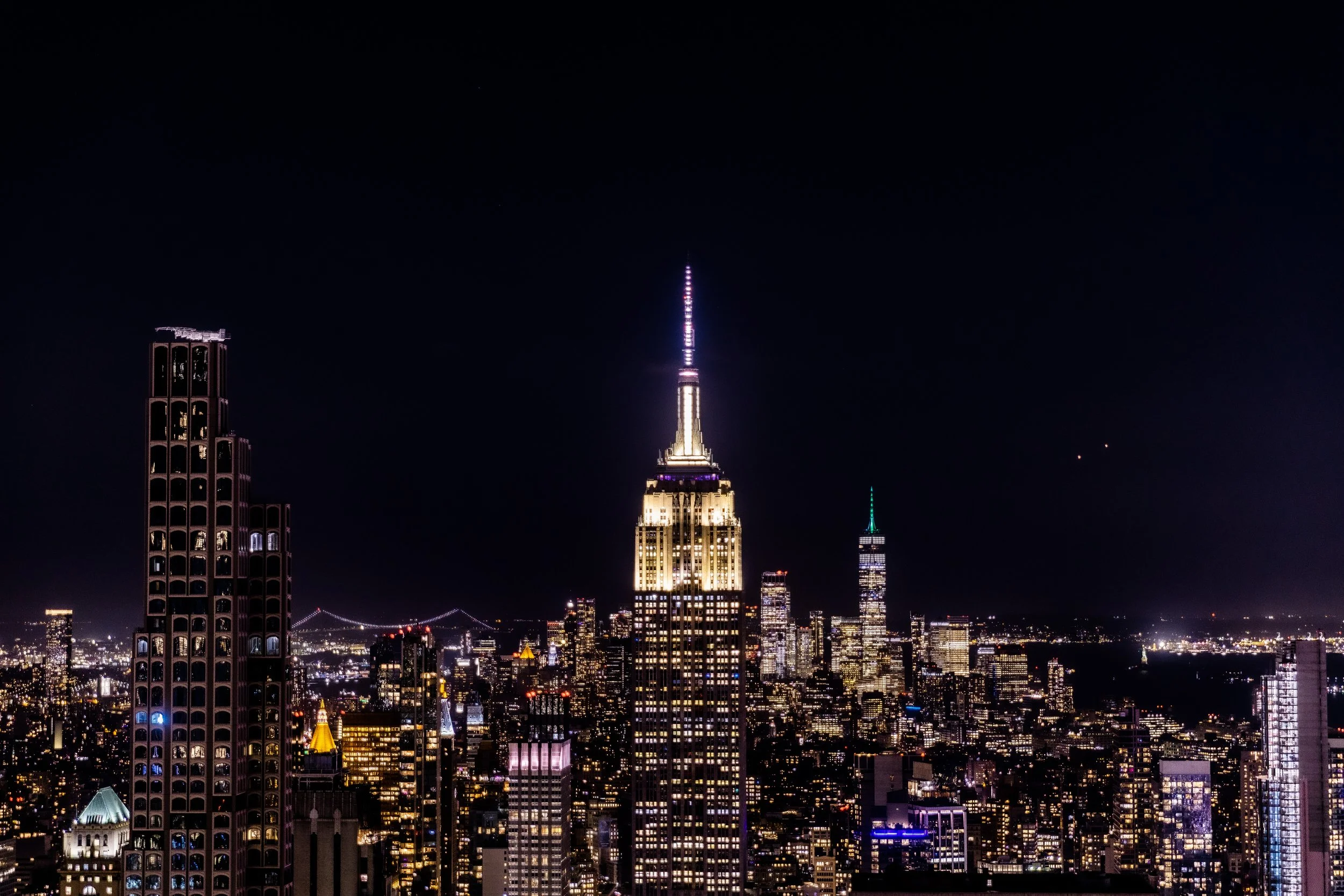 Nighttime cityscape of New York City with illuminated Empire State Building as the focal point, surrounded by other lit skyscrapers and a dark sky.