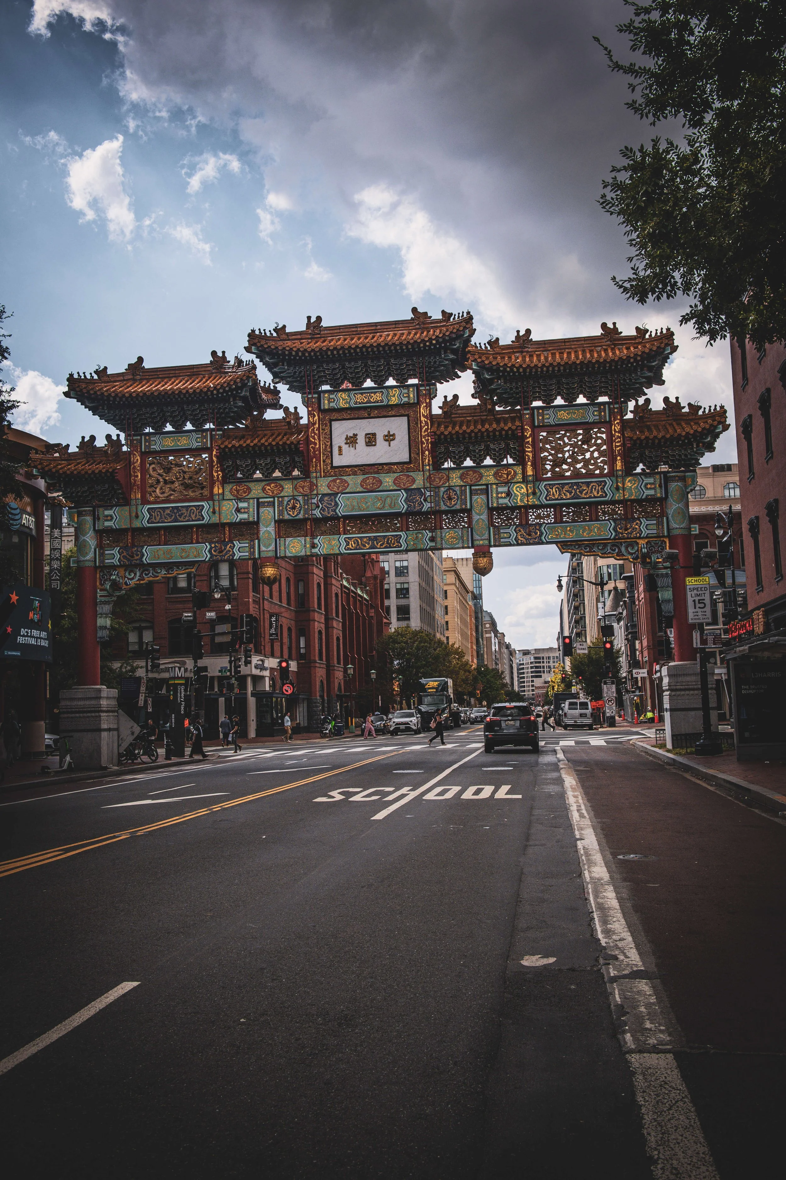 Urban street scene with a traditional Chinese style archway spanning the road, tall buildings on either side, and people walking across the crosswalk under a partly cloudy sky.