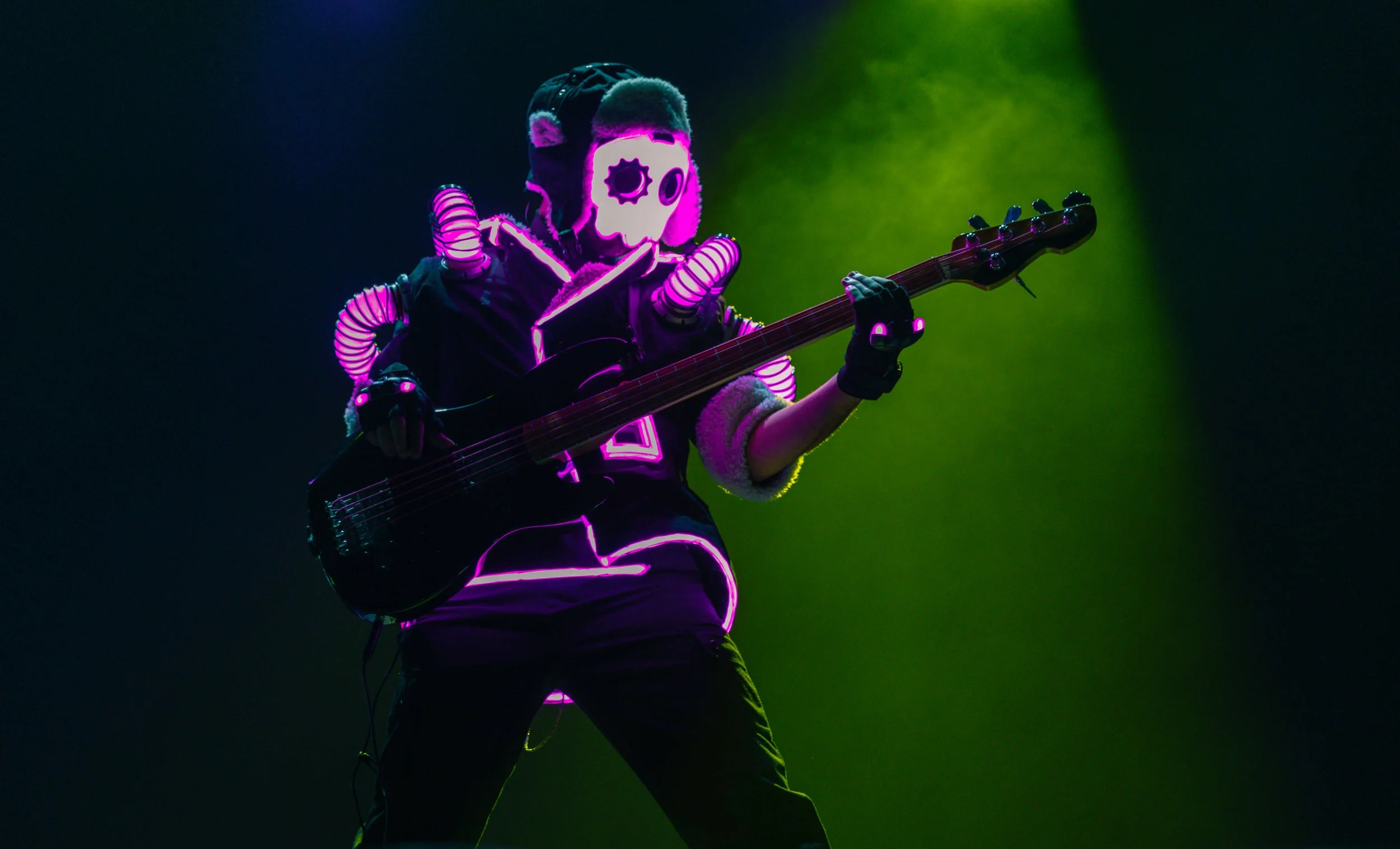 A musician wearing a skeleton mask with glowing neon accents, playing an electric bass guitar on stage with colorful lighting.