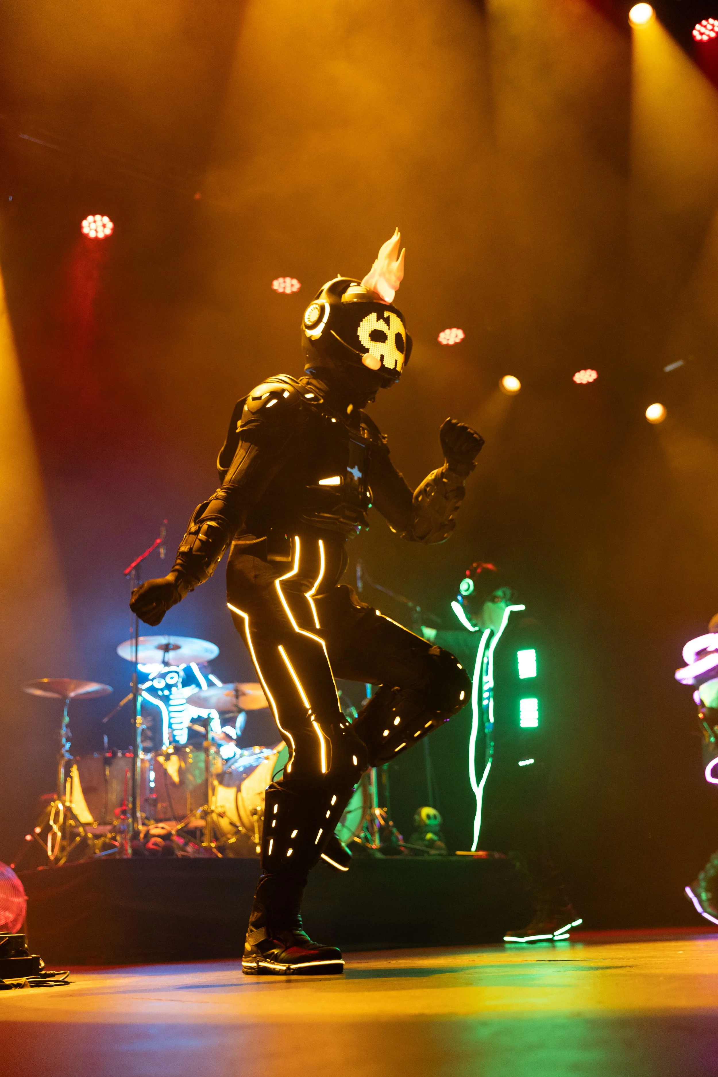 A performer dressed in black with neon piping is dancing on stage. The performer is wearing a costume with a skull mask and fire-shaped headgear. Bright stage lights and colorful neon decorations are visible in the background.