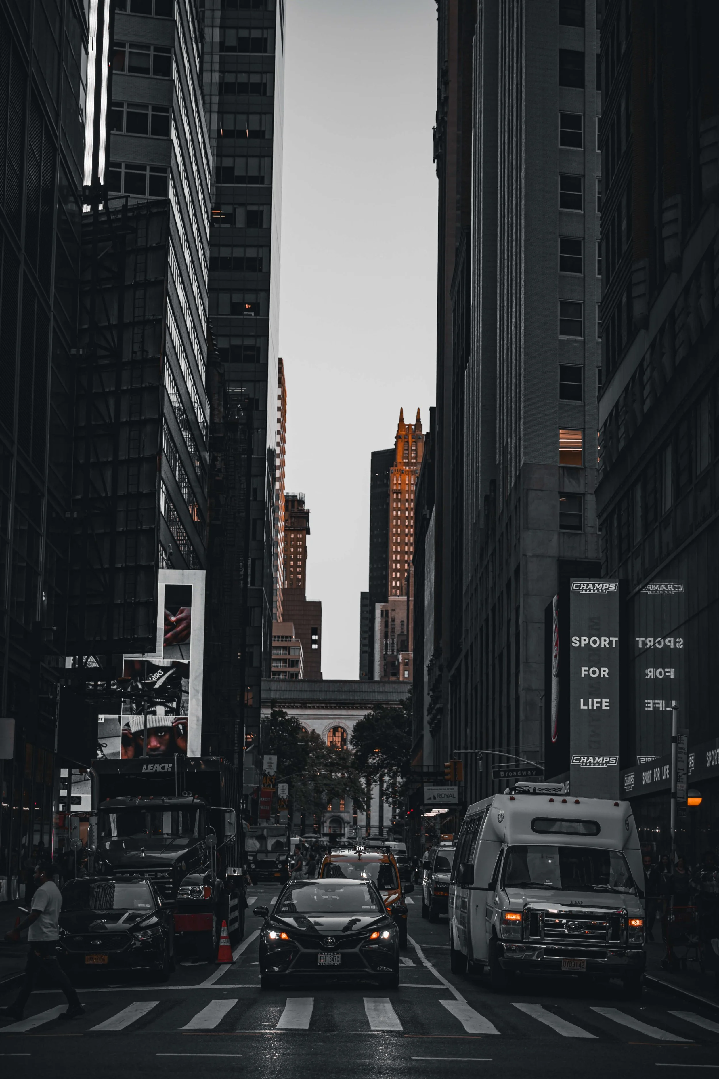 A city street in New York City with tall skyscrapers, cars, a van, pedestrians, and digital billboards, with an arched building in the distance.
