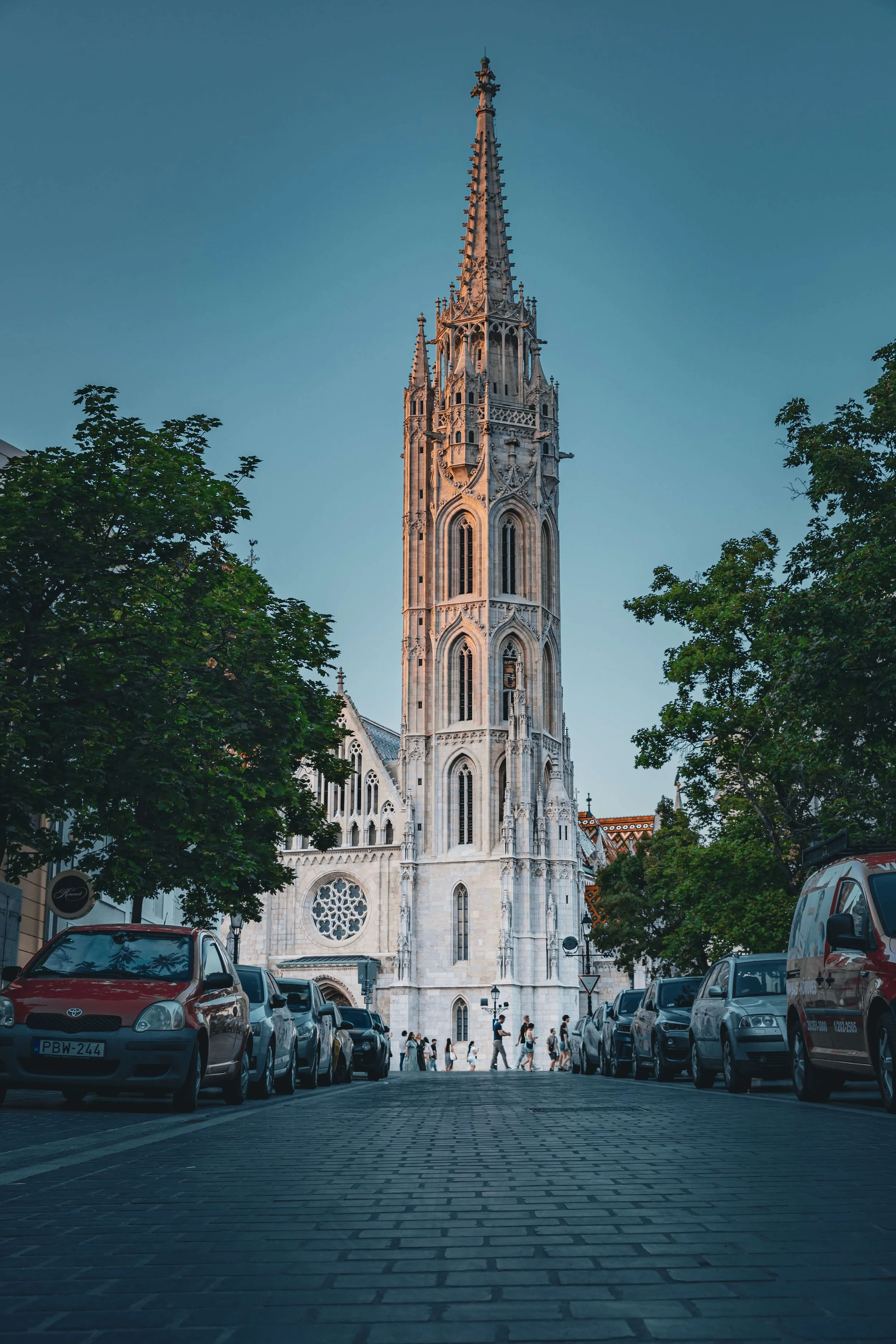 A tall Gothic-style church spire rising above a street with cars parked on either side and people walking across the street, with trees framing the scene.