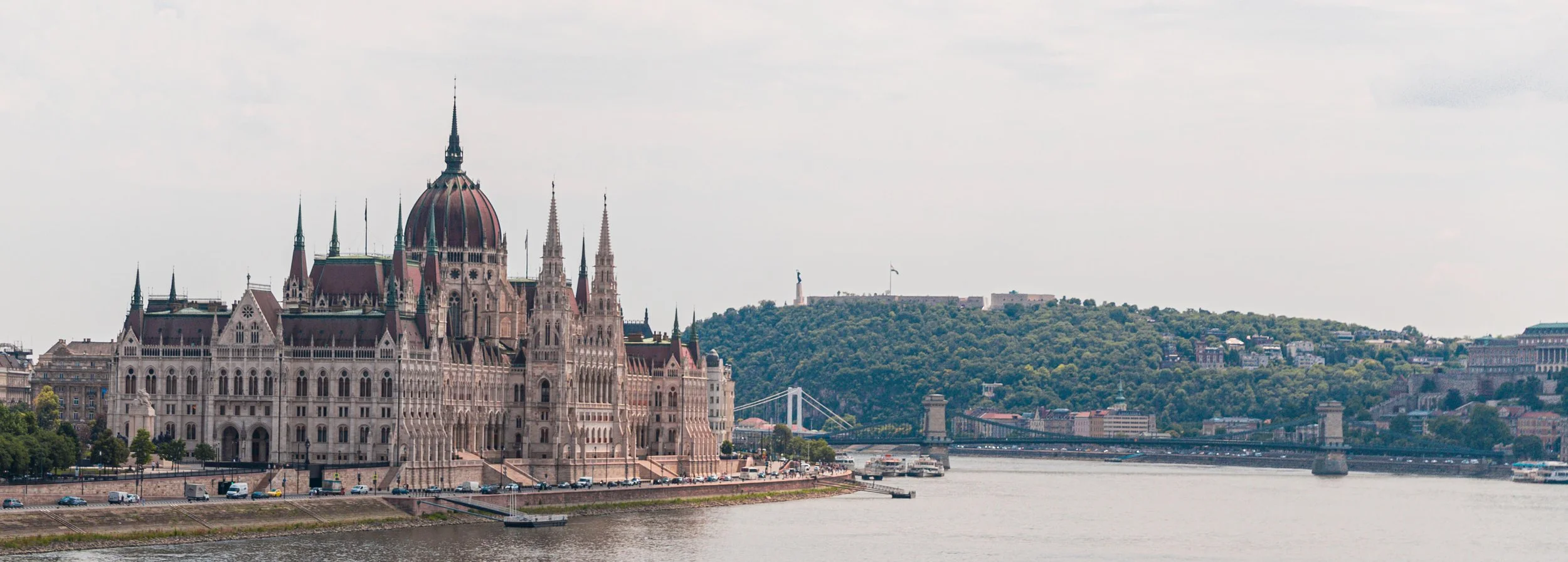 View of the Hungarian Parliament Building along the Danube River with a hill in the background that has a statue and flag.