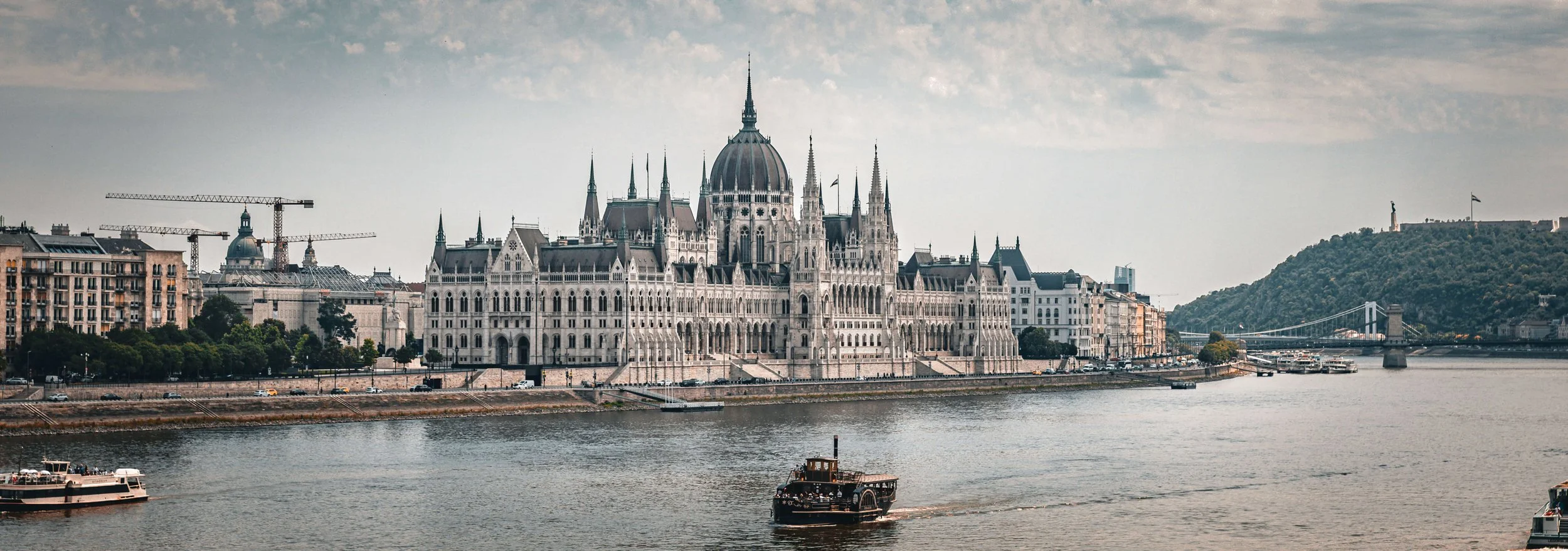 View of the Hungarian Parliament Building along the Danube River in Budapest, with boats on the water and a hill with a statue in the background.