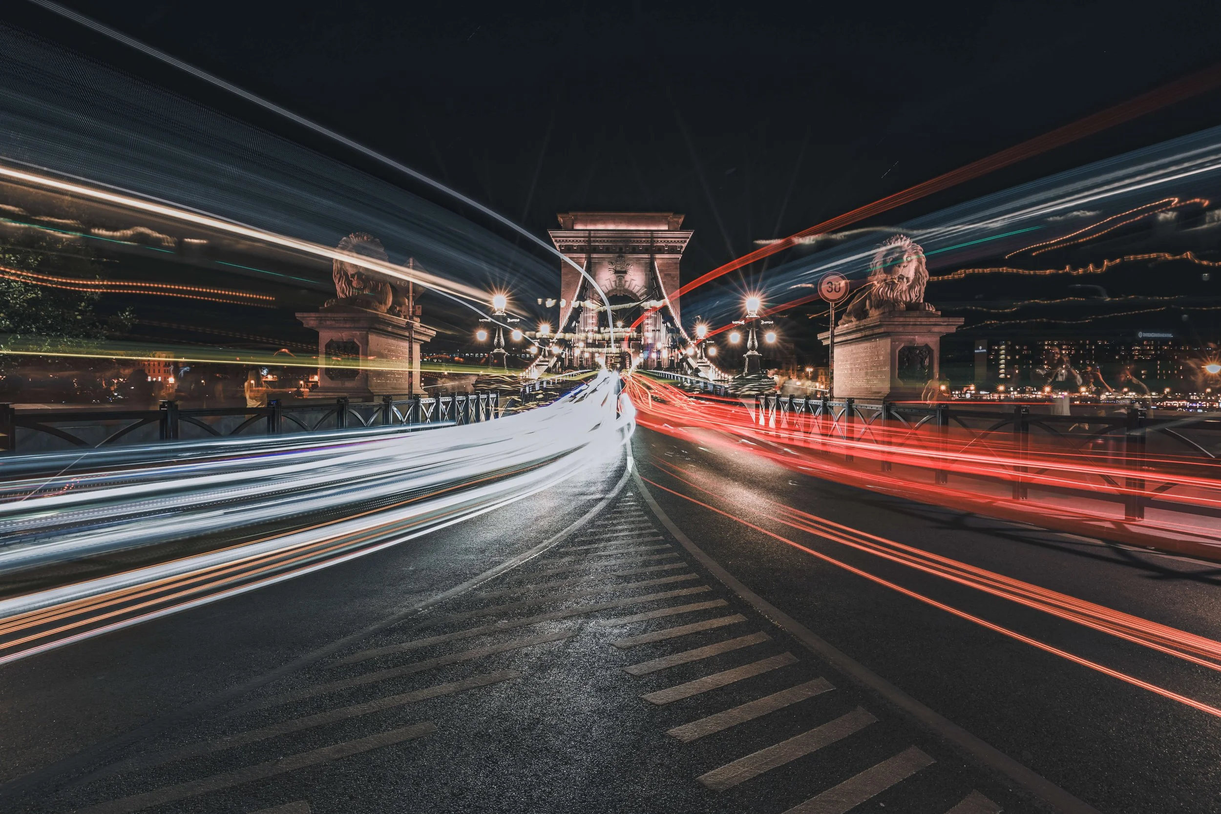 Nighttime photo of Chain Bridge in Budapest, Hungary, with light trails from passing vehicles, decorative lion statues at the bridge's entrance, and city lights in the background.