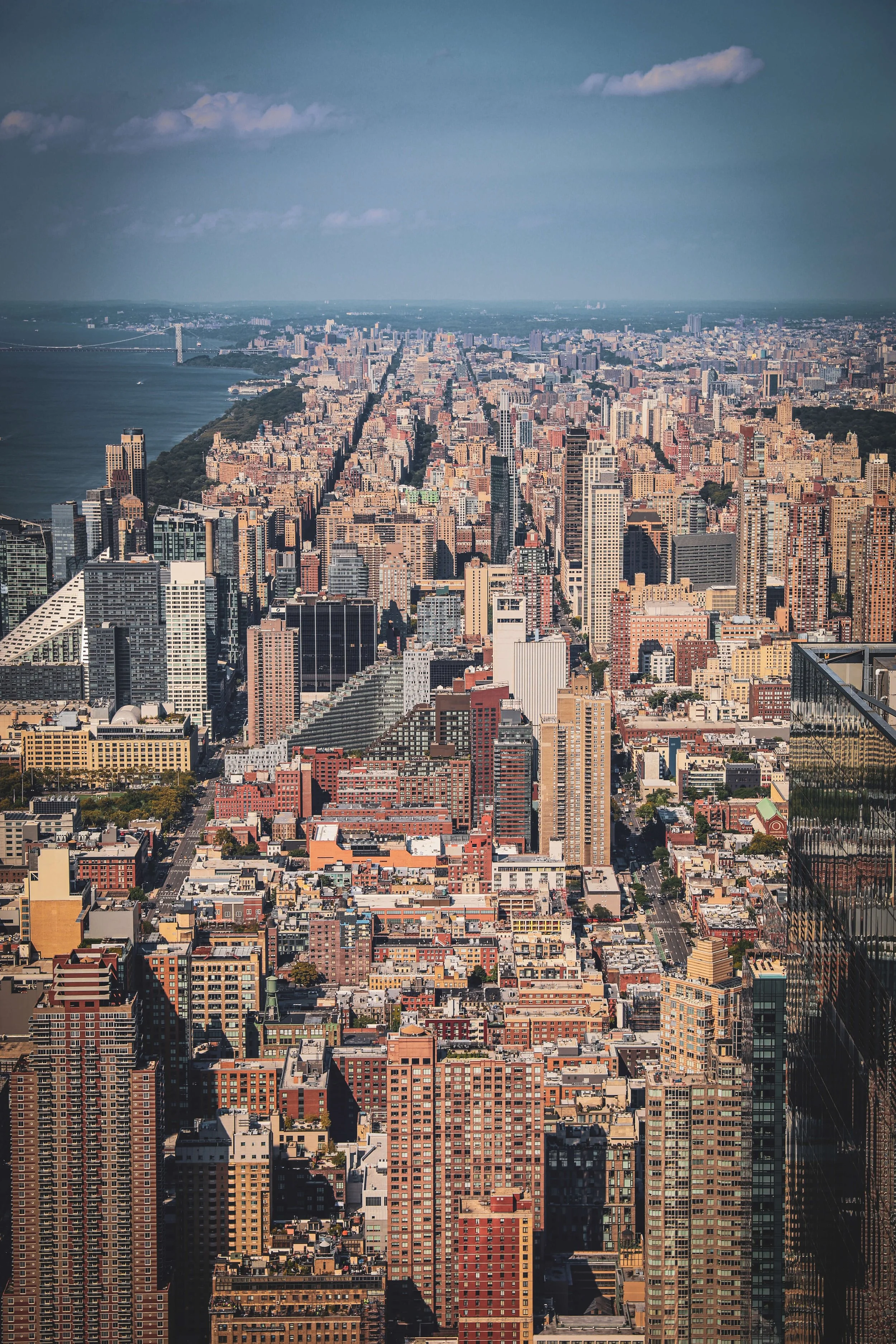 Aerial view of a densely populated cityscape with numerous skyscrapers, buildings, and streets, extending towards a river and distant horizon, possibly New York City.