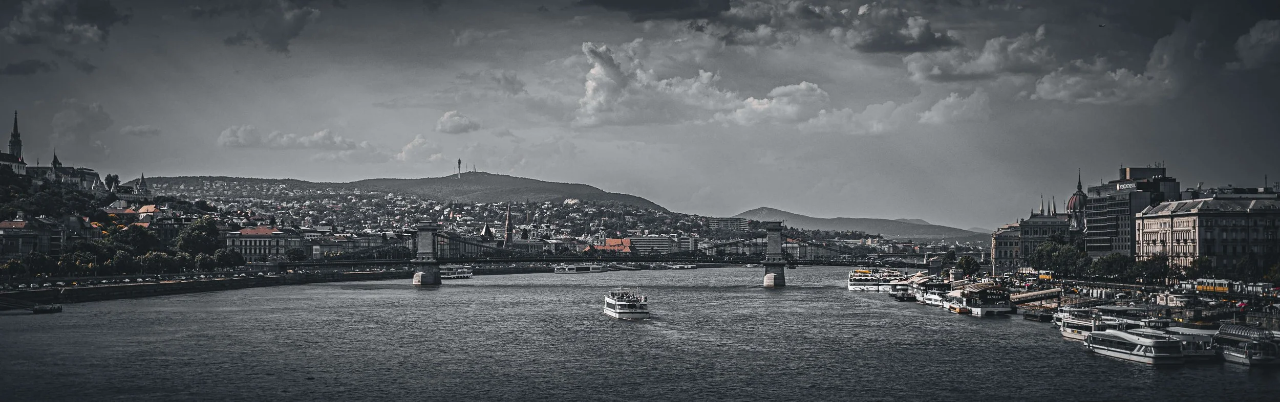 A black-and-white image of a cityscape with a river in the foreground, boats on the water, and a bridge crossing the river. The city has numerous buildings, some with spires, and hills in the background under a cloudy sky.
