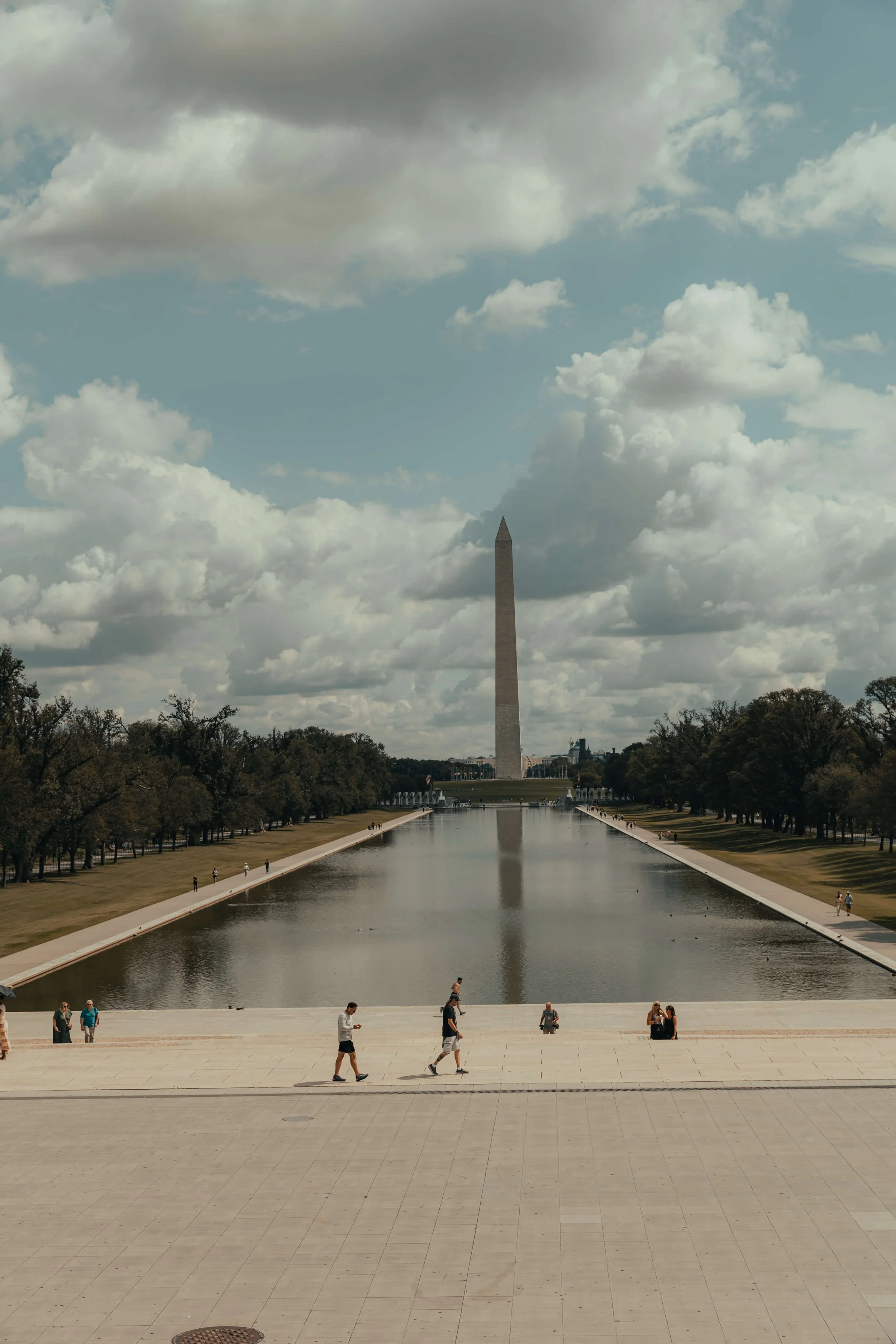 Washington Monument viewed from the Lincoln Memorial with the Reflecting Pool in the foreground, cloudy sky above, and several people walking and sitting around.