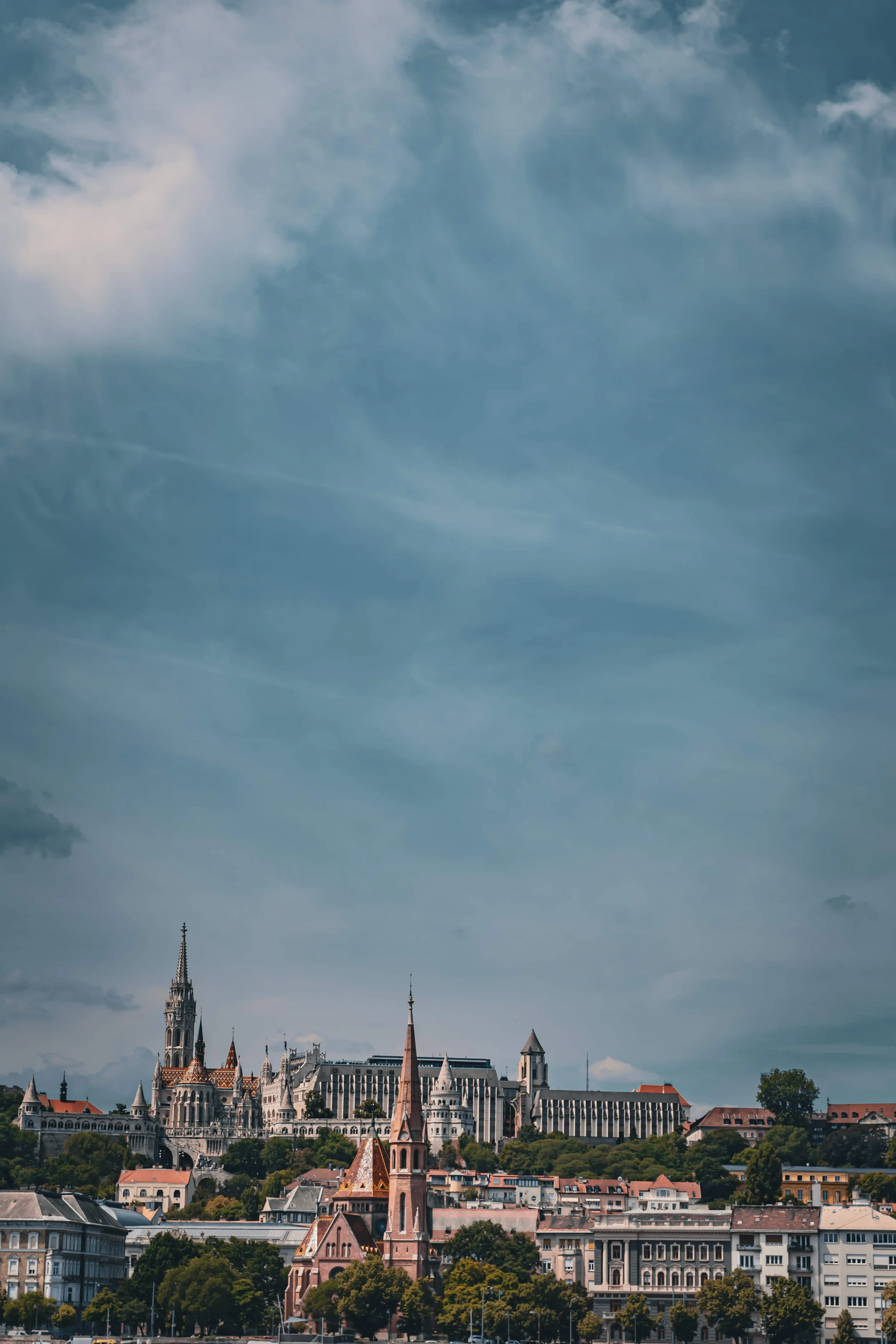 City skyline with historic buildings, church steeples, and green trees under a cloudy sky.