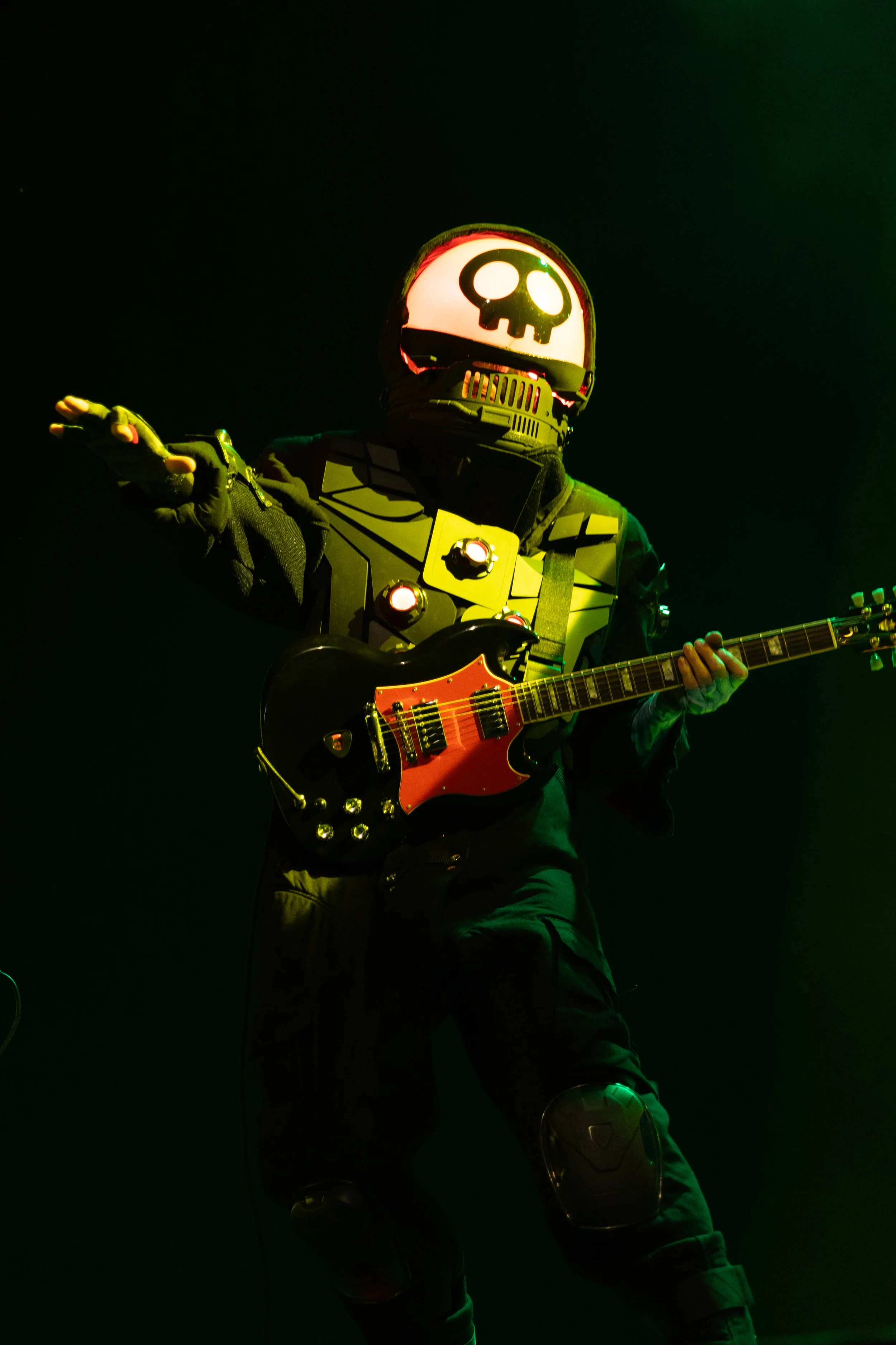 A person in a robotic suit with a skull face mask playing an electric guitar on a dark stage with green lighting.