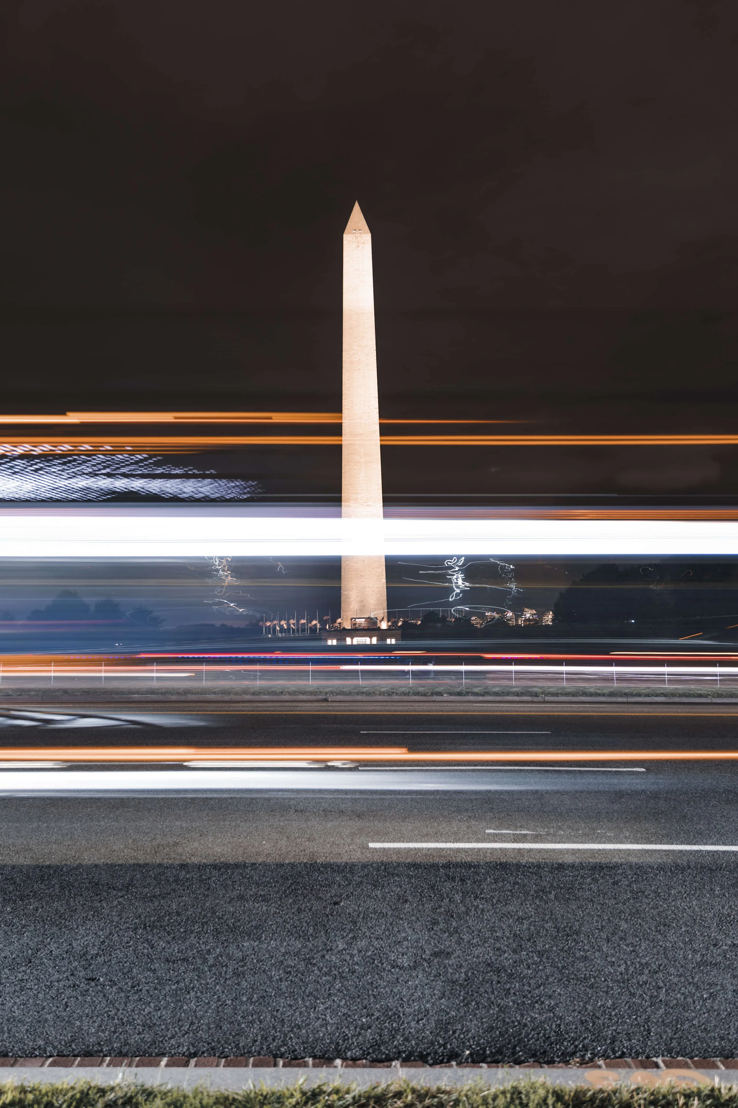 Nighttime view of the Washington Monument in Washington, D.C., with light trails from passing vehicles in the foreground.