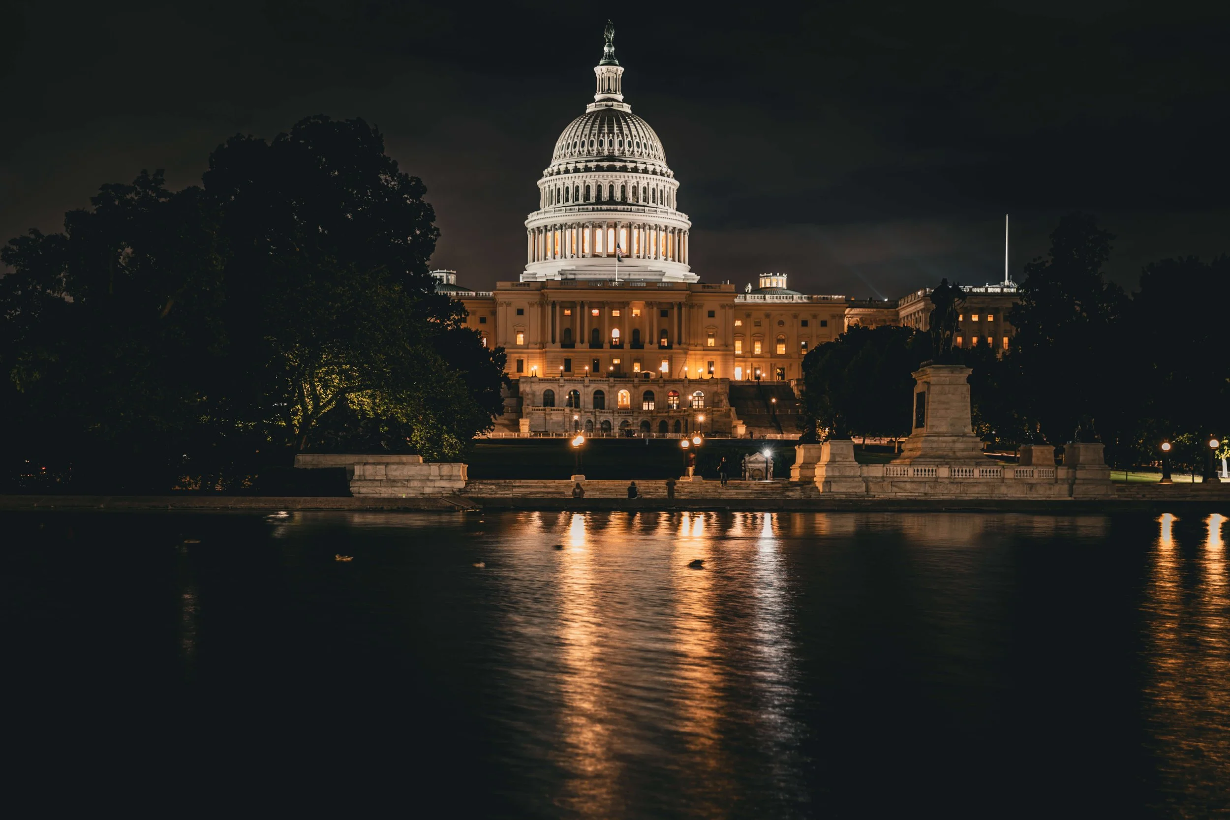 Night view of the illuminated U.S. Capitol building in Washington, D.C., with reflections on the water in the foreground and trees surrounding the area.