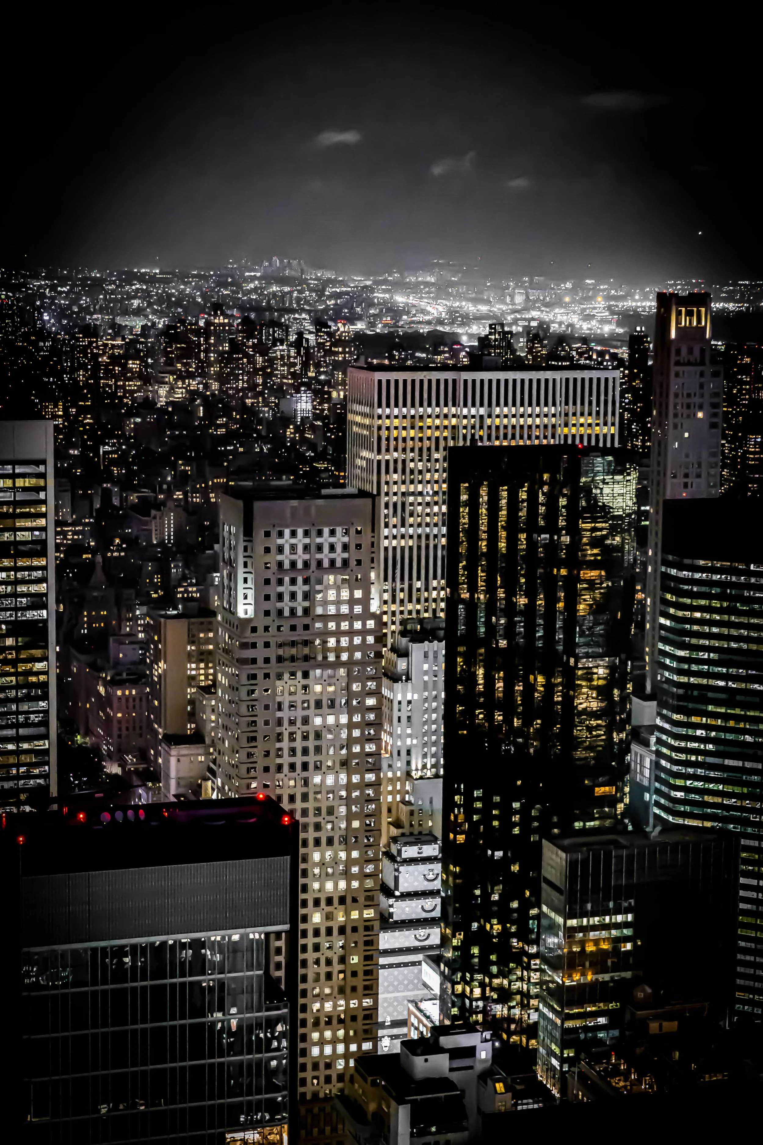 Nighttime aerial view of a cityscape with illuminated skyscrapers and buildings, with a faint view of the city stretching to the horizon.