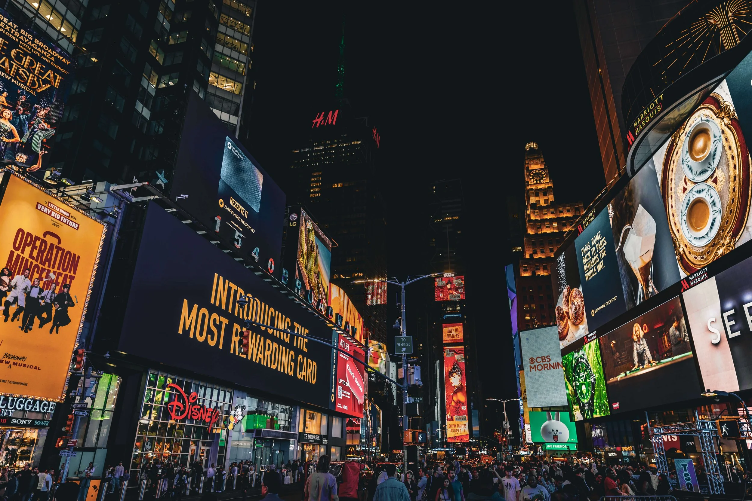 Brightly lit billboards in Times Square at night with a crowd of people walking and standing below.