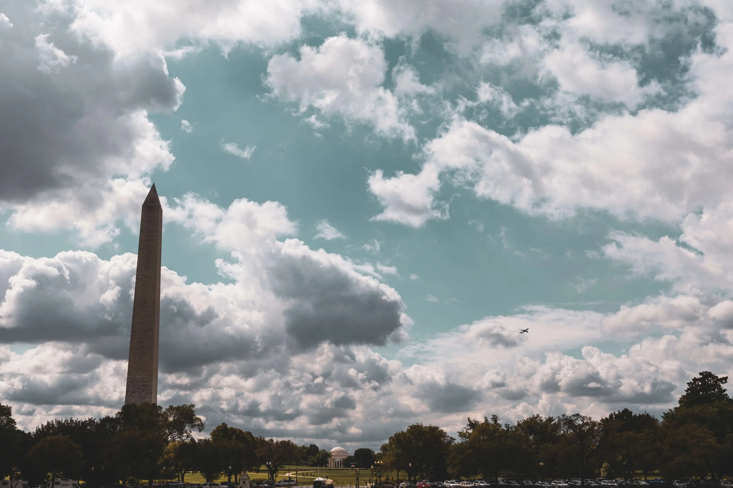 Washington Monument under a partly cloudy sky with trees and parked cars in the foreground