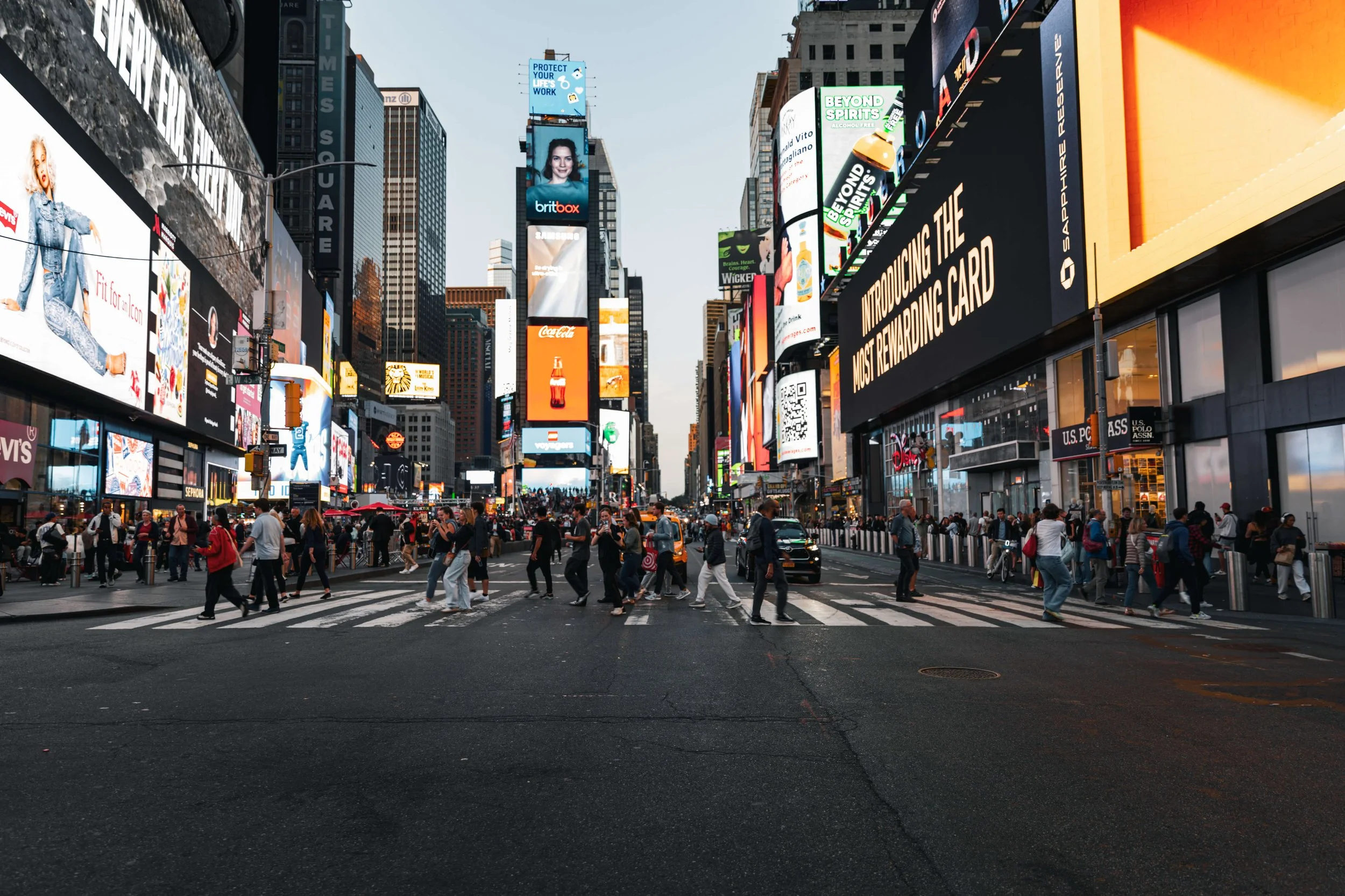 Crowded city street crossing in Times Square, New York City, with people walking, tall buildings with digital billboards, and advertisements for Coca-Cola, BritBox, Disney, and more.