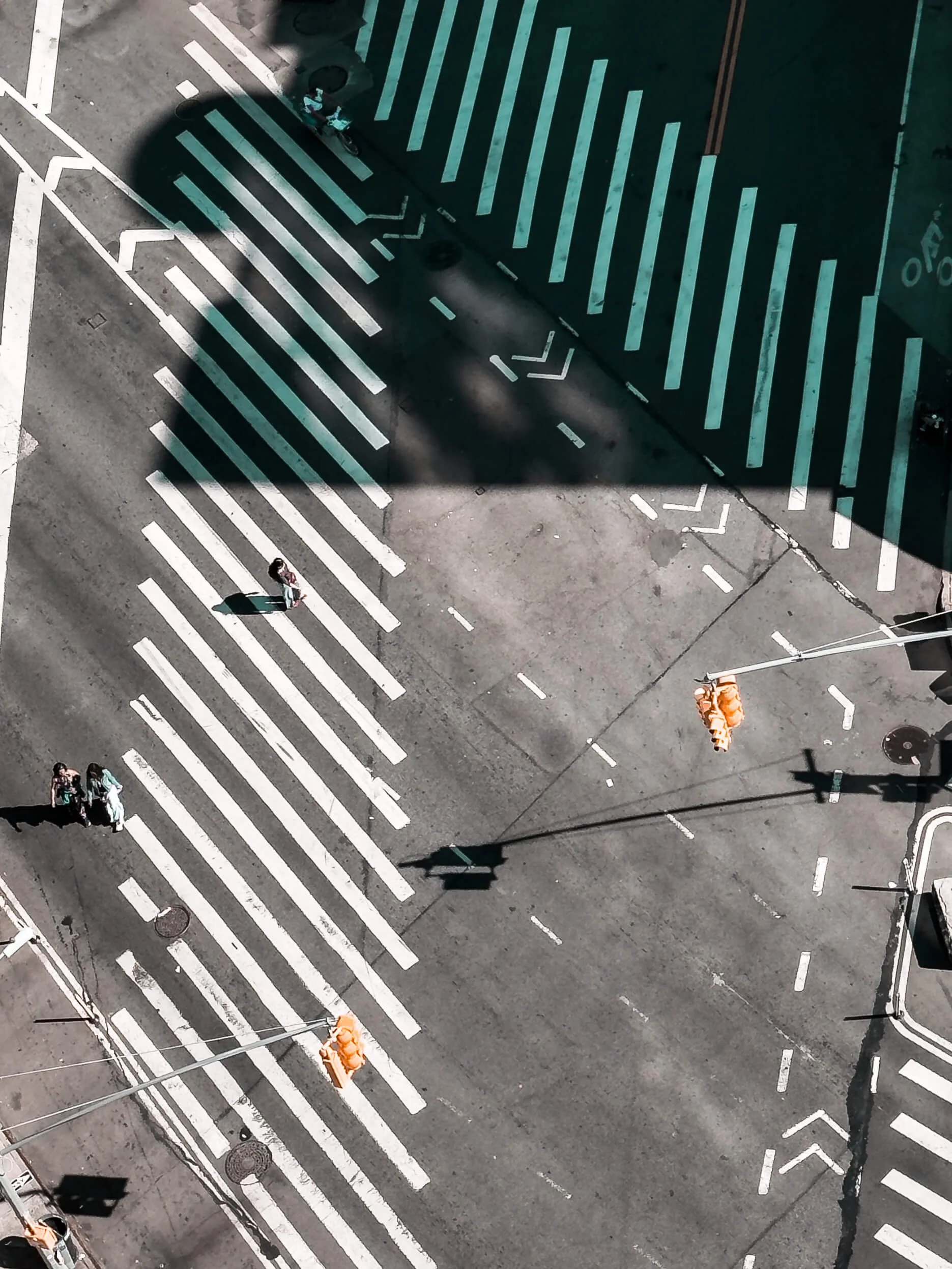 An aerial view of a city intersection with crosswalks, traffic lights, and three pedestrians walking across the street. A shadow of a building is cast over part of the crosswalks.