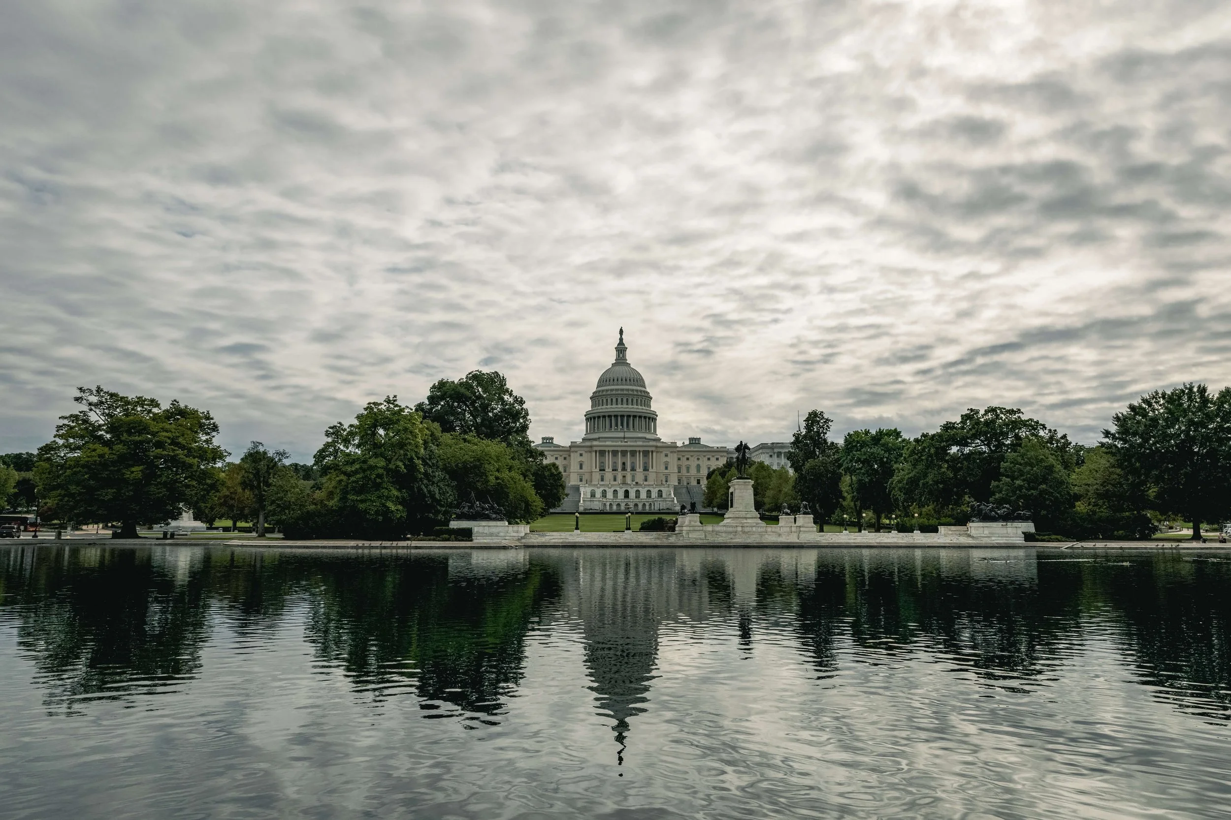 The United States Capitol building with its dome, trees surrounding it, and a pond in the foreground reflecting the building and cloudy sky.